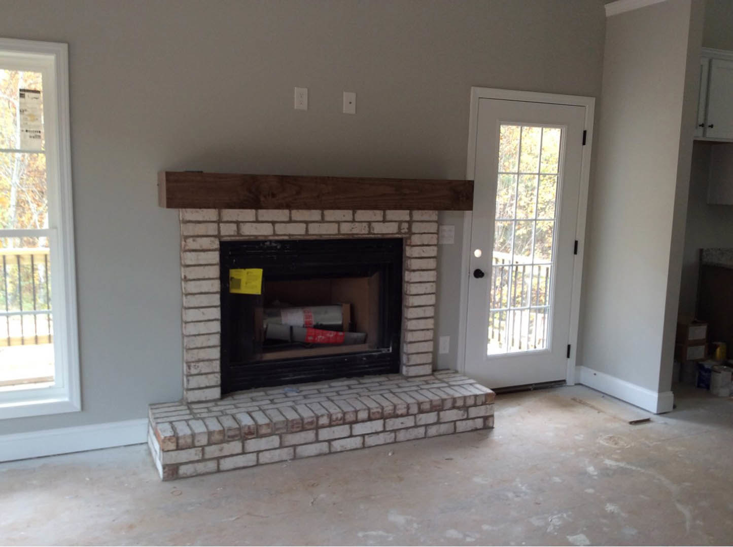 Brick fireplace with black fire screen, wood-burning stove, and hearth; box of yellow paper on dark surface nearby; adjacent door with window, large window with railing, and trees