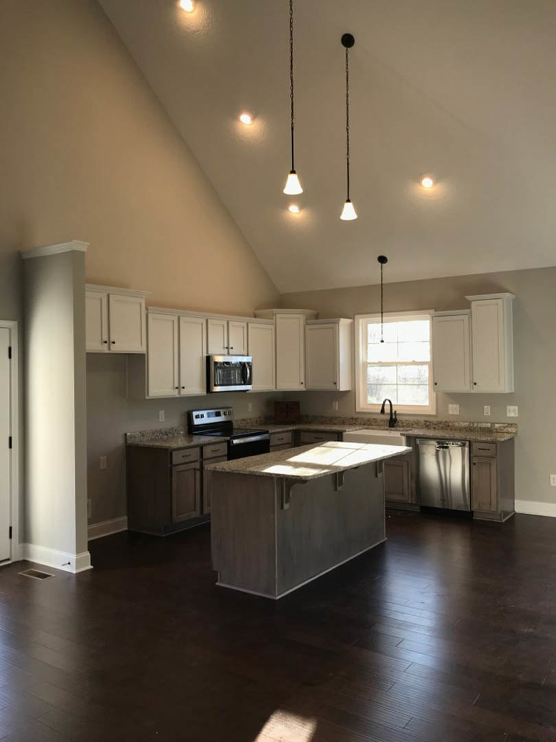 Spacious kitchen featuring a large ceiling, dark wood flooring, granite-topped island, white walls with black handles, built-in microwave, and sunlit window.