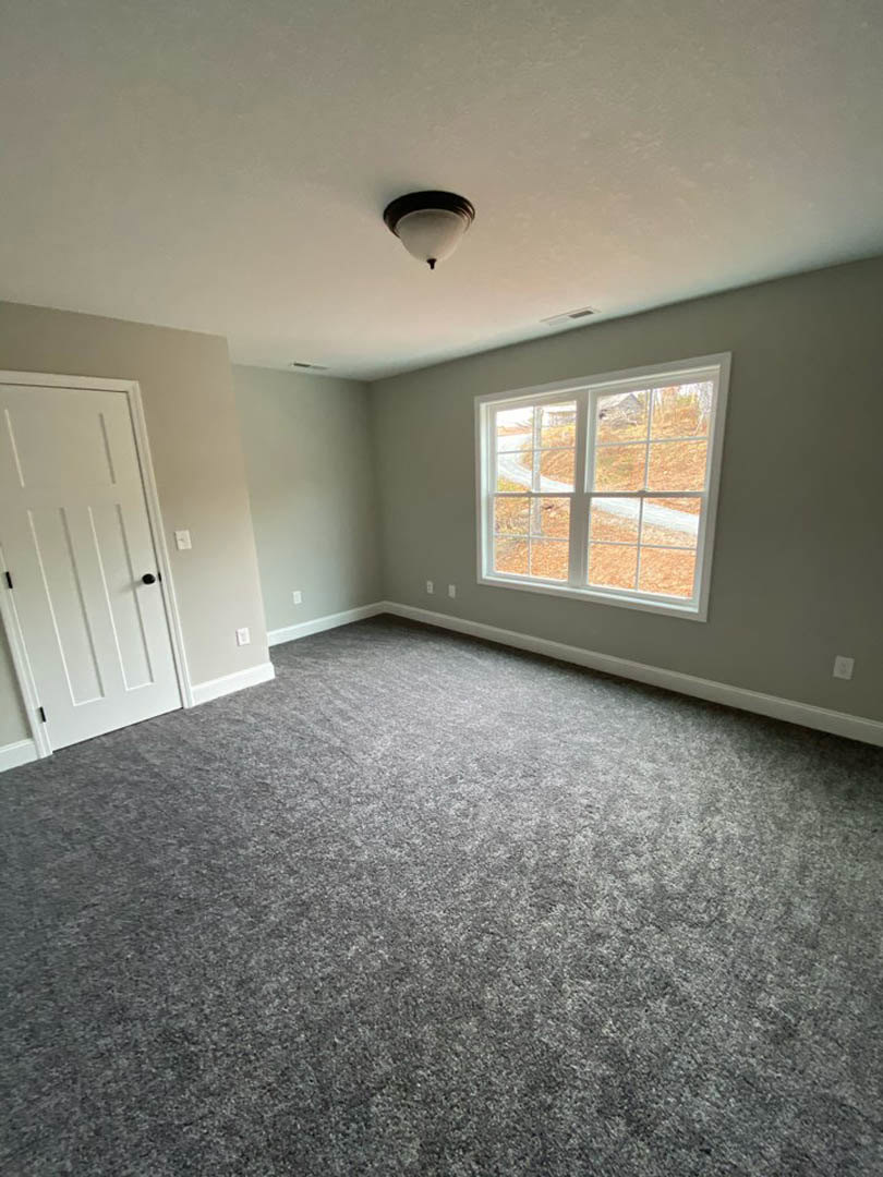 Grey carpeted room with white walls, large window showing autumn trees, white door with black knob, ceiling-mounted light fixture.