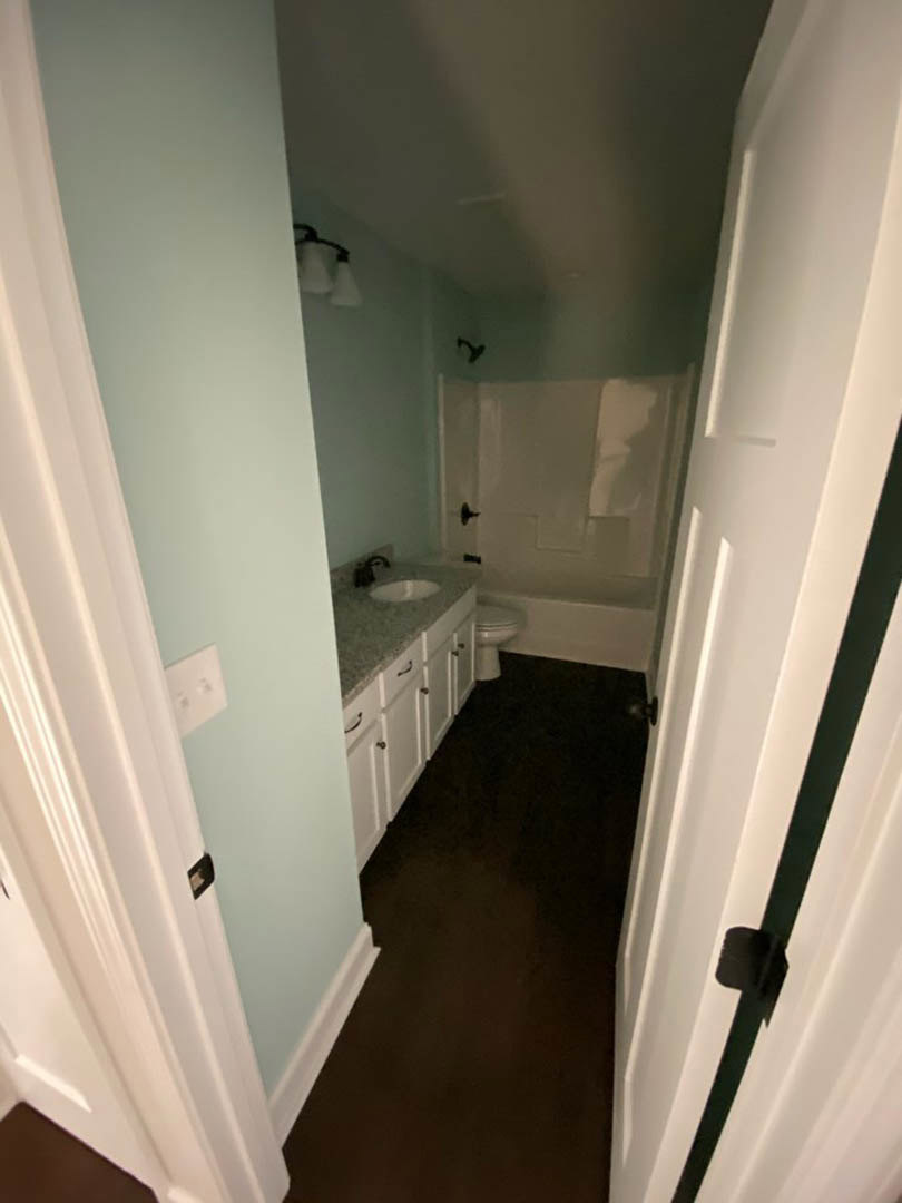 White bathroom featuring a pedestal sink, modern toilet, dark wood flooring with white trim, and a wall-mounted lamp with black base.