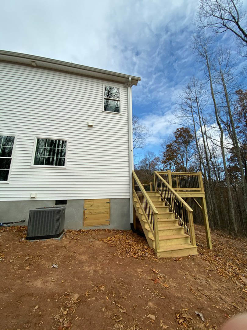 White siding house with large windows, elevated wooden deck, black metal railings, and staircase leading to landscaped yard; trees reflected in glass, cloudy sky above.