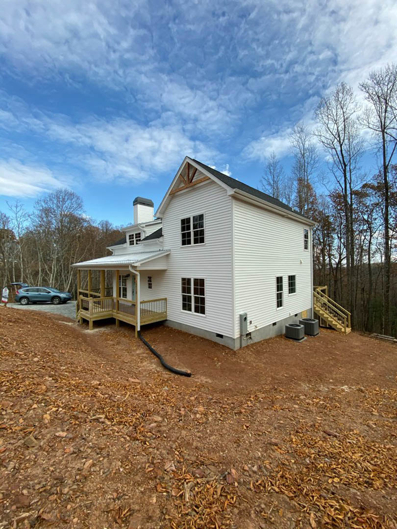 White cottage-style home with covered porch, wood deck, large windows, and a car parked behind the house; blue sky with scattered clouds and surrounding greenery.