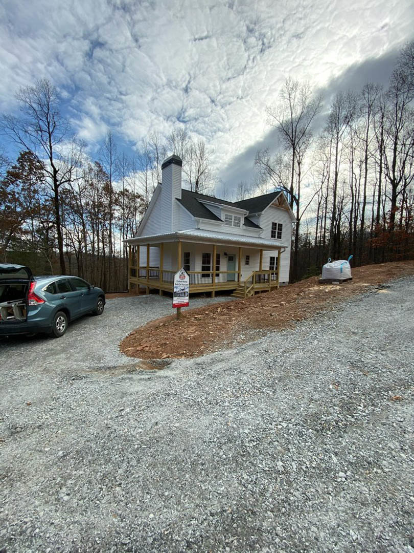 Gray siding house with covered porch, white columns, and a car parked in the driveway with trunk open; small sign on porch and white bag with blue straps near entrance; trees and