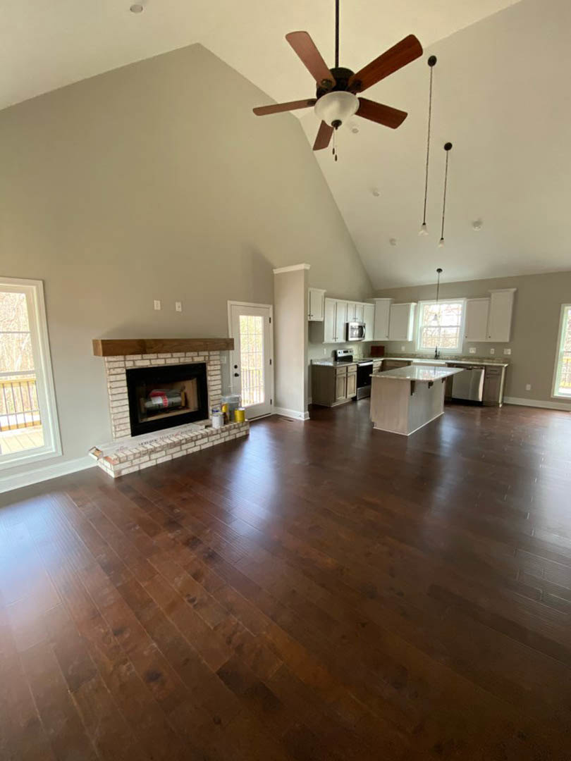 Spacious living room featuring wood flooring, stone fireplace with white mantel, ceiling fan with light fixture, large windows, and open view into kitchen with white marble