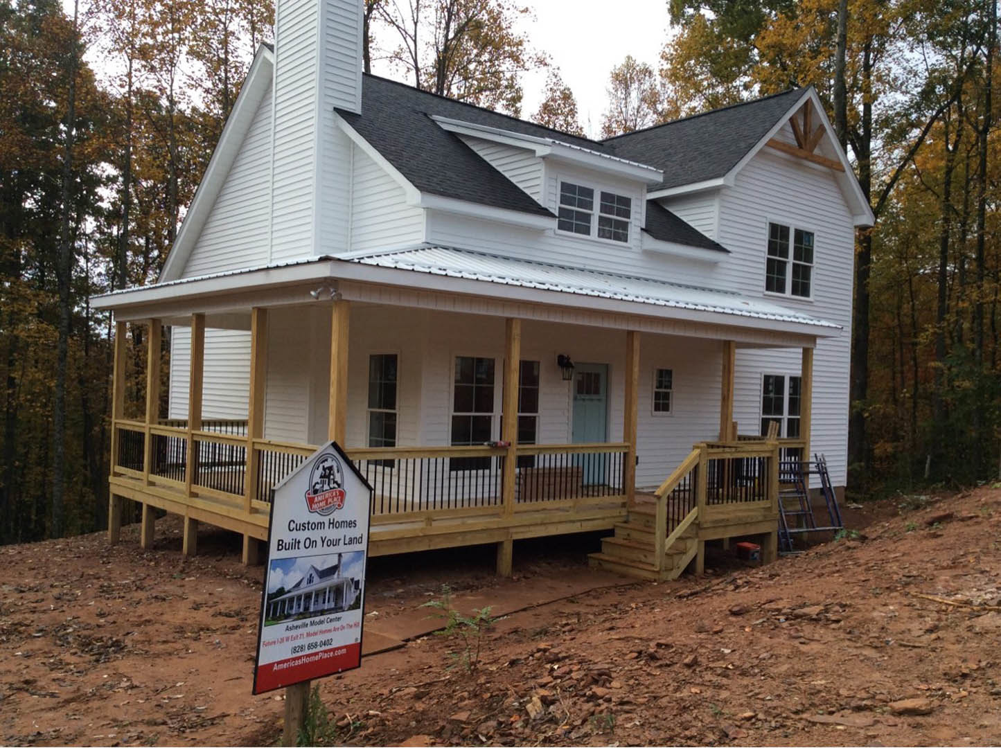 Two-story house with light siding, white-framed windows, wooden front porch with railing, sign displayed near entry, small plant growing beside dirt path, leafy tree in background