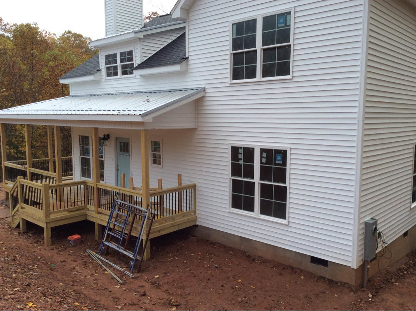 Two-story house with light siding, elevated wooden deck, staircase leading to yard, large windows, and sloped roof; ladder leaning against deck railing, plastic storage container