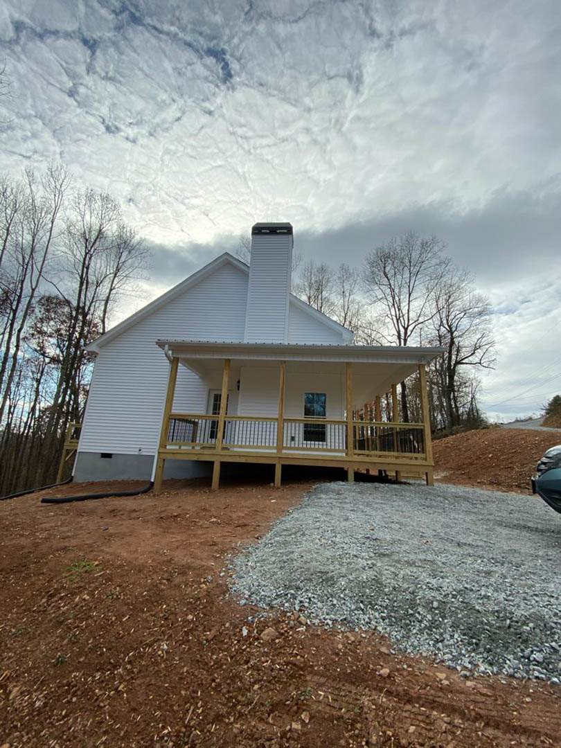 White siding house with covered front porch, gravel driveway, double-hung windows, and mature trees in the background
