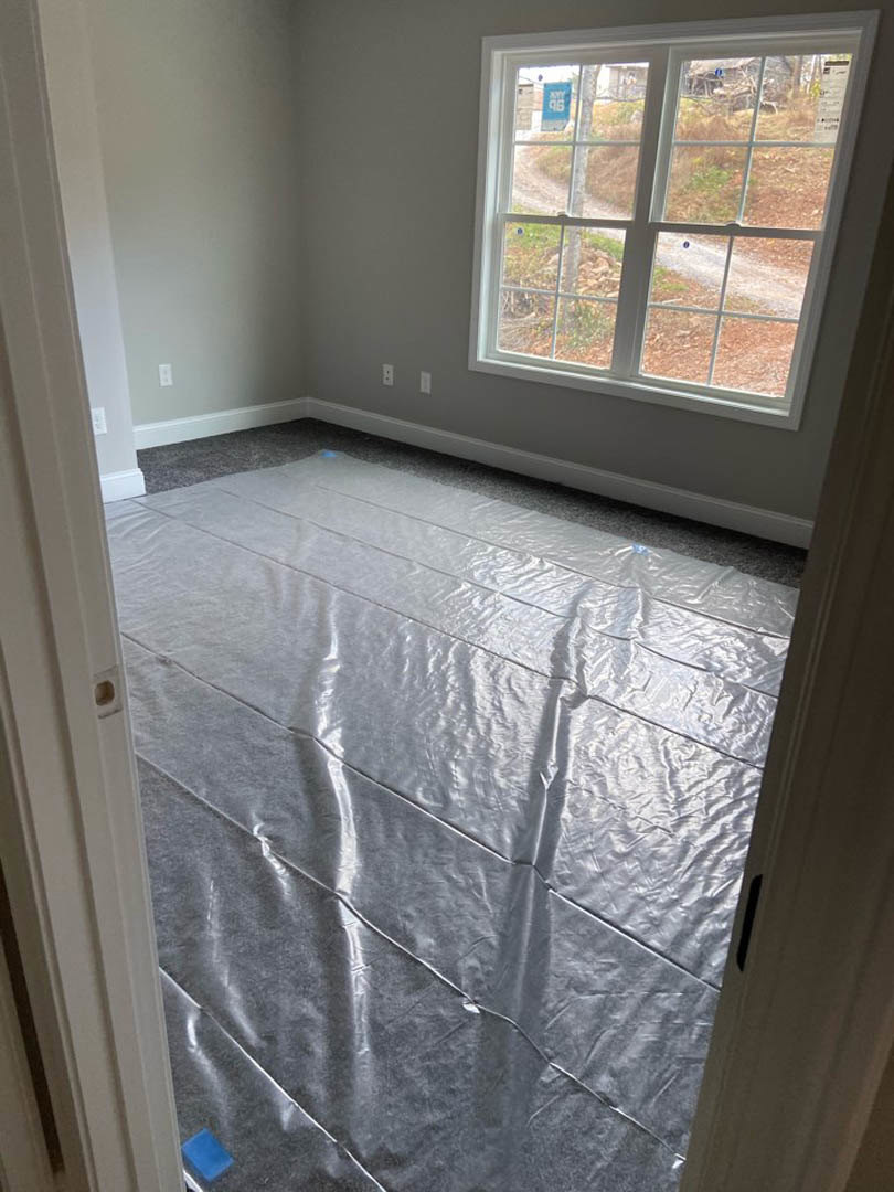 Unfinished room with white plaster walls, plastic sheeting covering the floor, large window overlooking dirt path, close-up of white door, natural daylight illuminating interior.