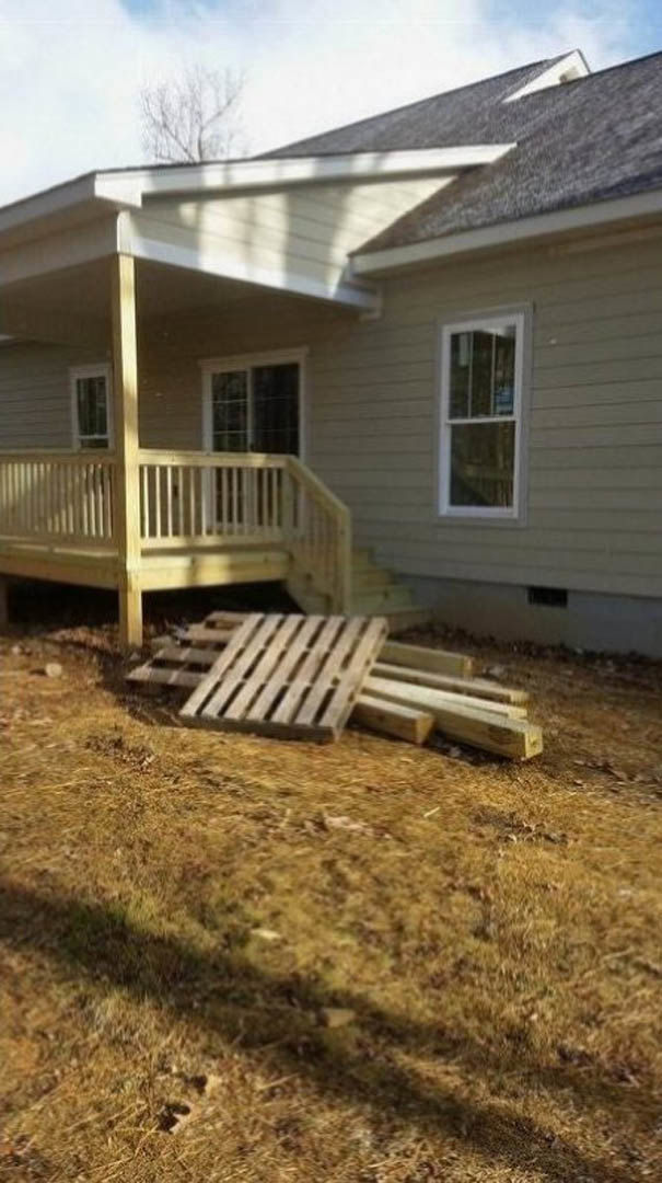 Wooden pallets stacked on grass beside a house with white-framed windows, light siding, and a covered porch.