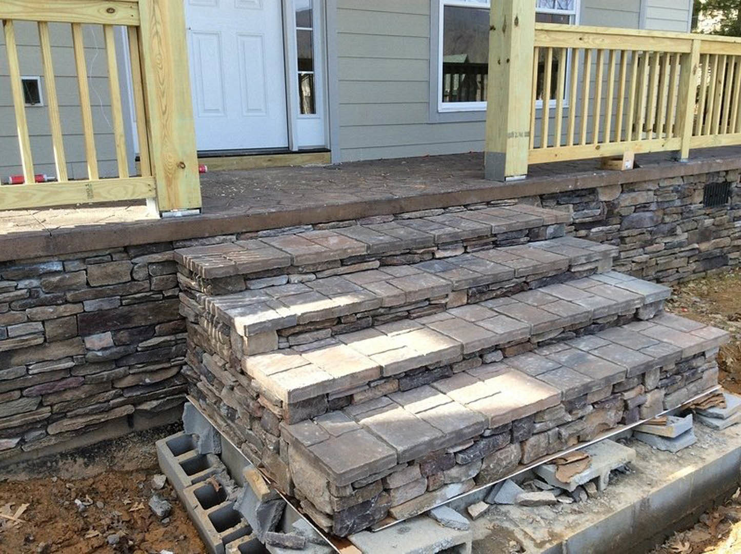 Stone steps leading up to a porch with a wooden fence, white wall, and window with railing in the background