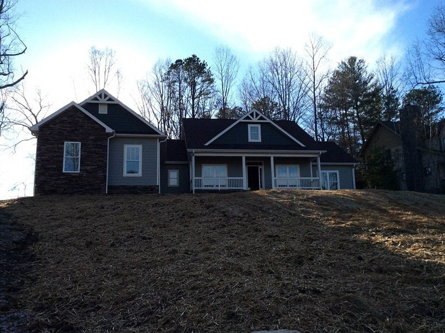 Two-story brick house with white-framed windows and white railing, situated on a grassy hill with trees and cloudy sky in the background