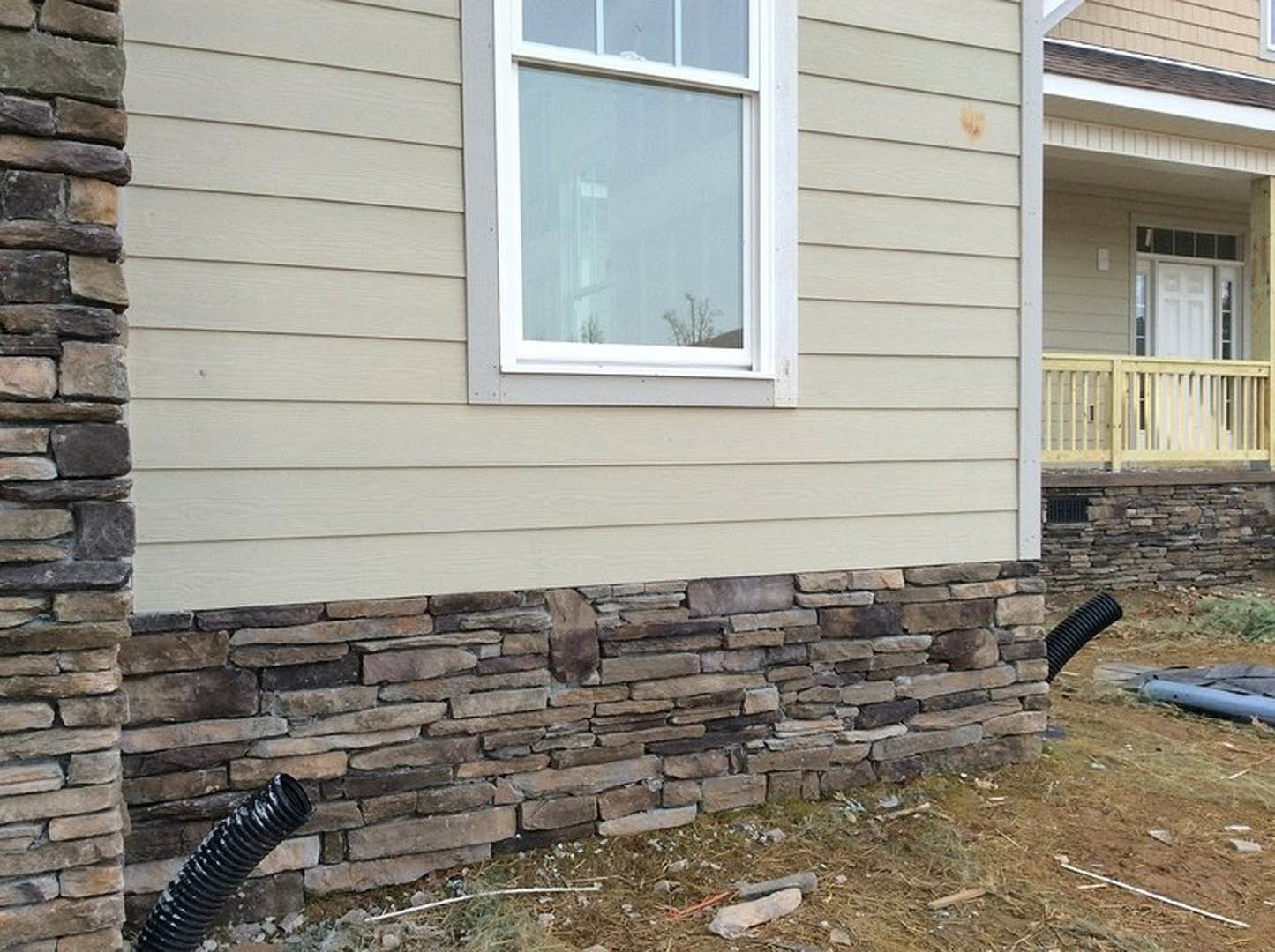 Red brick exterior with white-trimmed window, wooden railing, black pipe resting on grass near stone wall, scattered debris visible