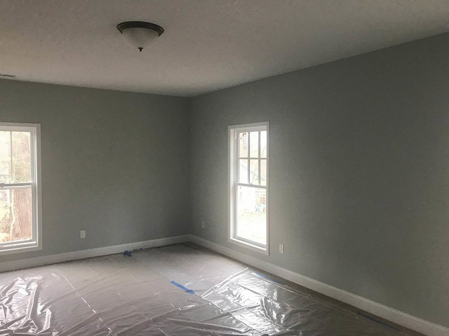 White-framed window set in plastered wall, plastic sheet covering light-colored floor, blue painter’s tape and black object resting on surface, natural daylight illuminating