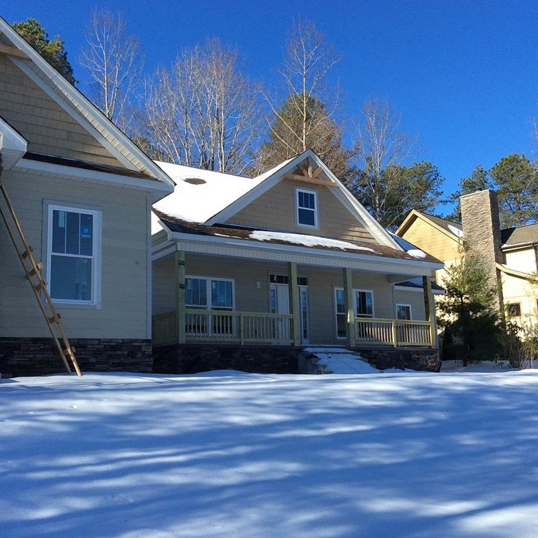 Snow-covered yard with bare trees, neighboring houses in the background, white-framed window with a sign, ladder leaning against exterior wall, snow blanketing roof and ground with
