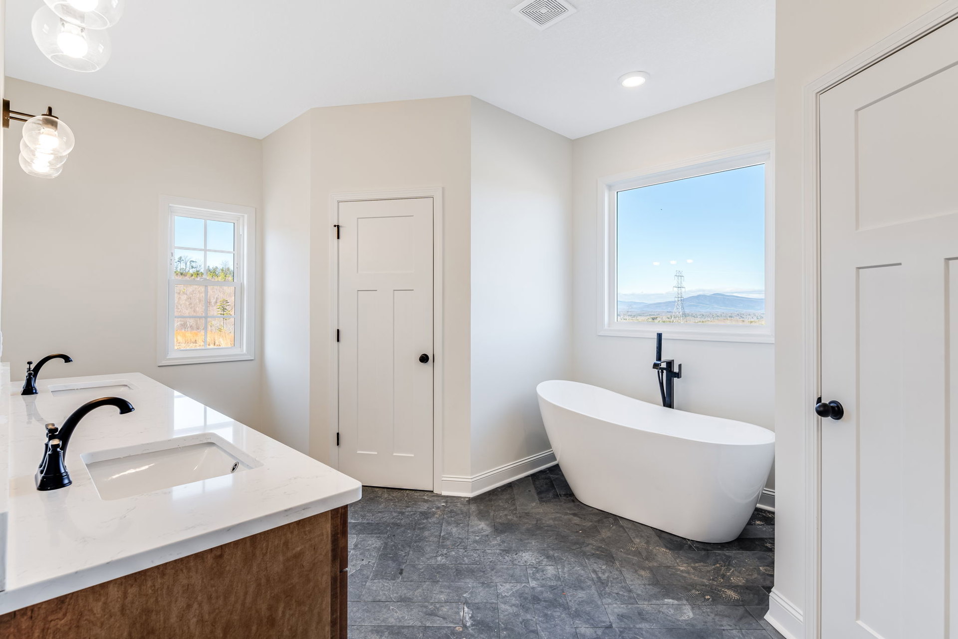 Bathroom with white freestanding tub featuring black exposed plumbing, white sink and faucet, tiled floor, window overlooking trees and grass, white door with black hardware