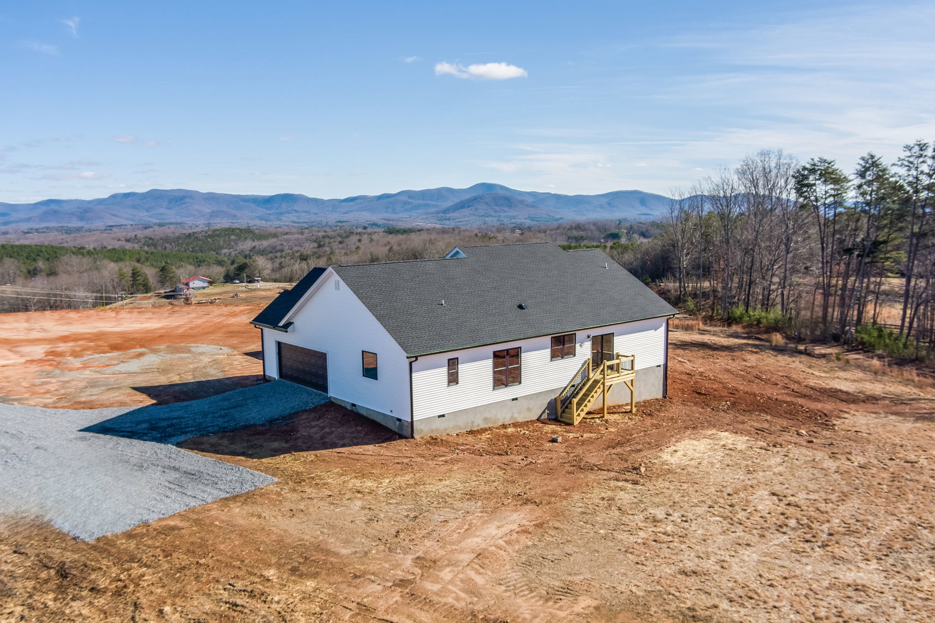 Modern house with black roof, white exterior walls, wooden staircase leading to entrance, large window, situated in a dirt field under a partly cloudy blue sky