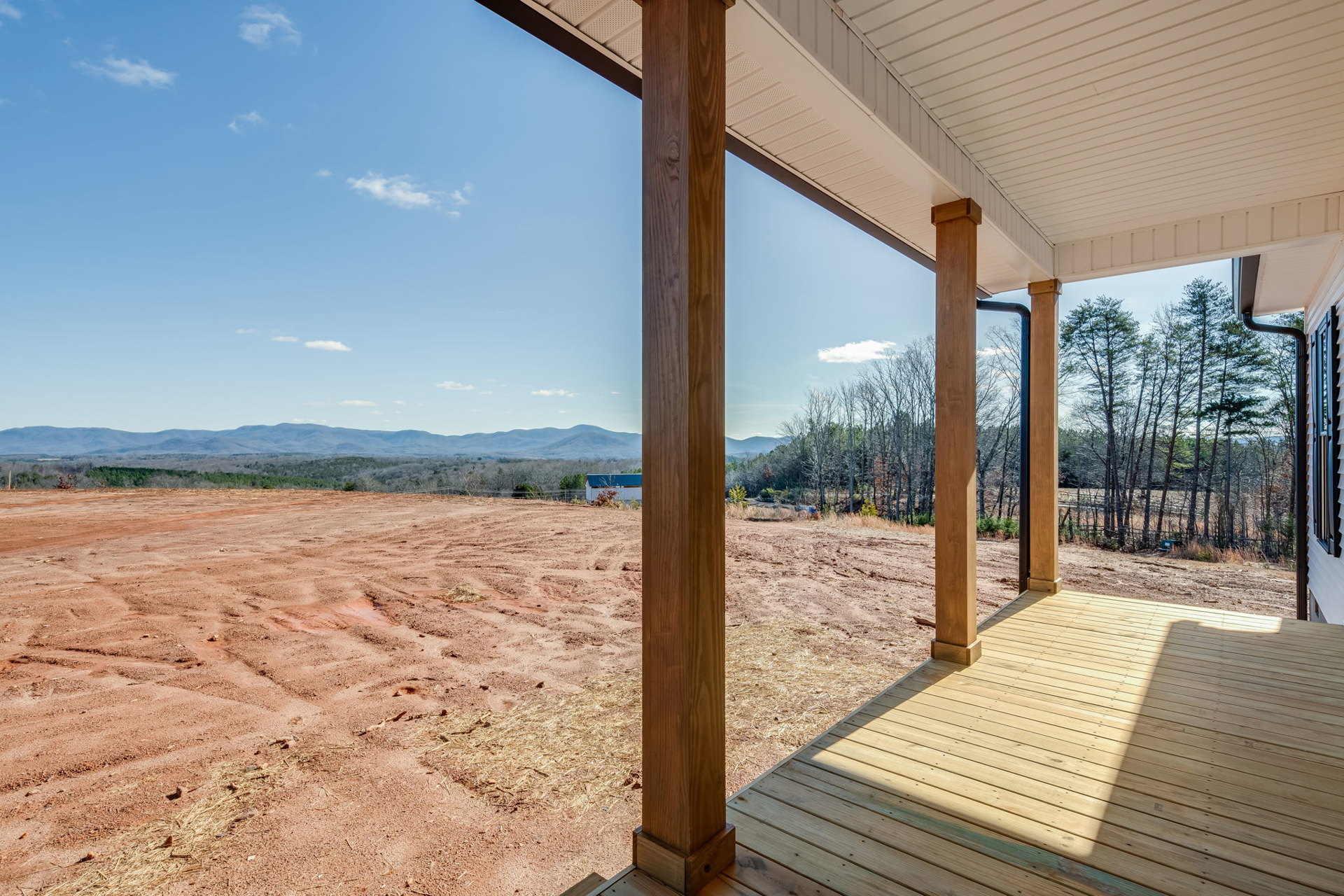 Wooden porch with white roof and pillars overlooking expansive dirt field, grassy area and trees in background under partly cloudy sky