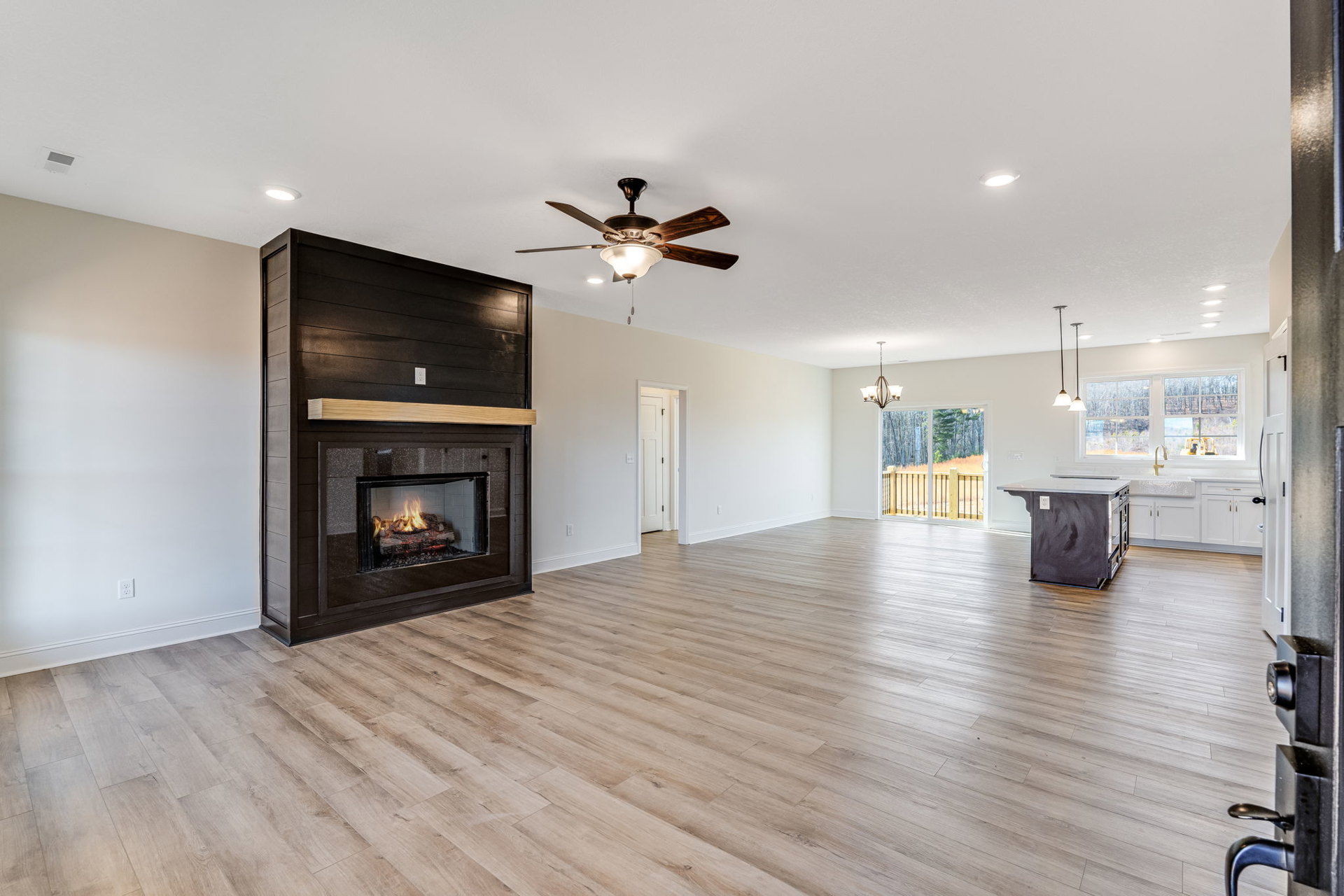 Living room with hardwood flooring, stone fireplace with wood mantel, ceiling fan with light fixture, partial view of kitchen island with drawer
