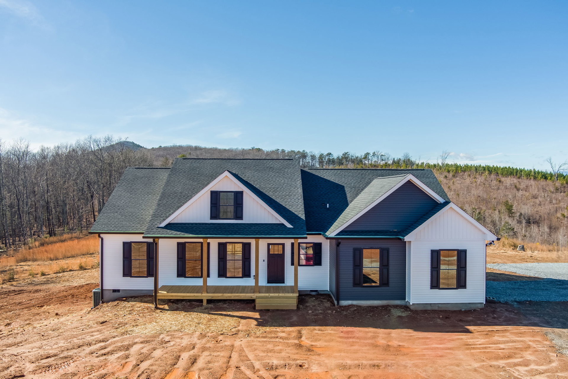 Two-story house under construction with exposed framing, dark brown door featuring a square window, glass-paneled door, porch area, blue shutters on windows, surrounded by trees