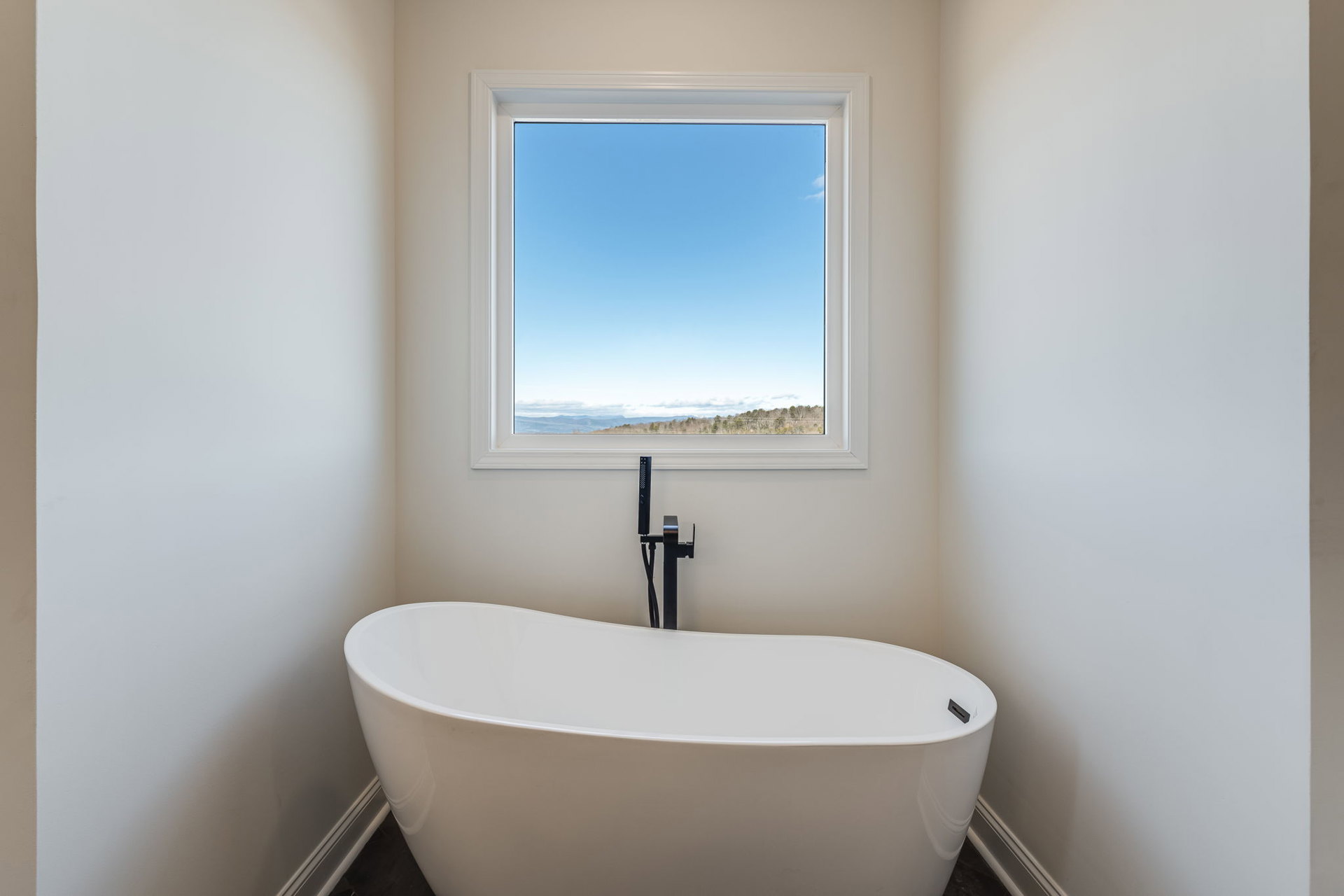Freestanding white bathtub beside large window with mountain view, white walls, chrome faucet, and minimal decor