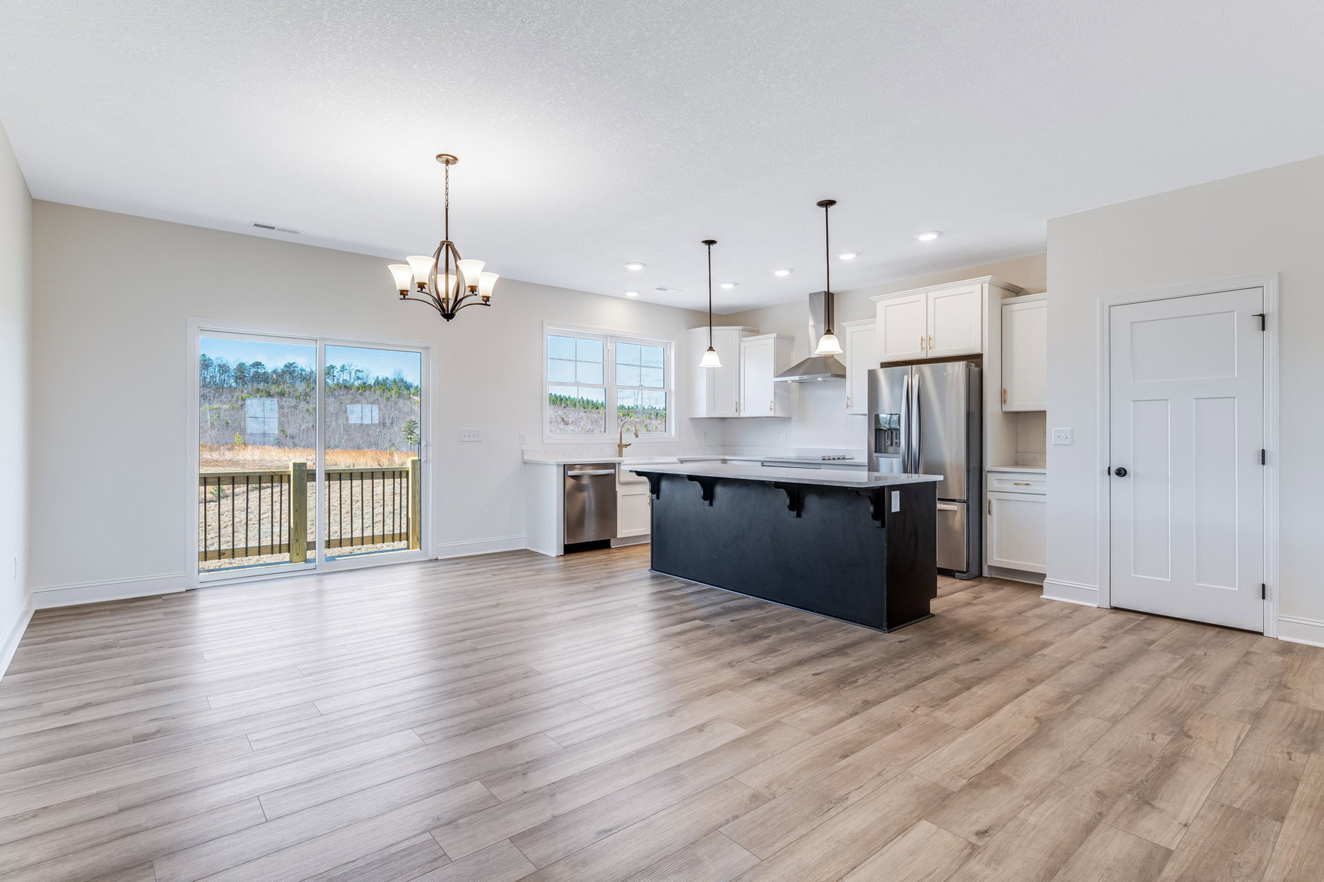 Kitchen with large black island topped in white, stainless steel refrigerator, wood flooring, black accent wall, sliding glass door opening to fenced yard and trees, white