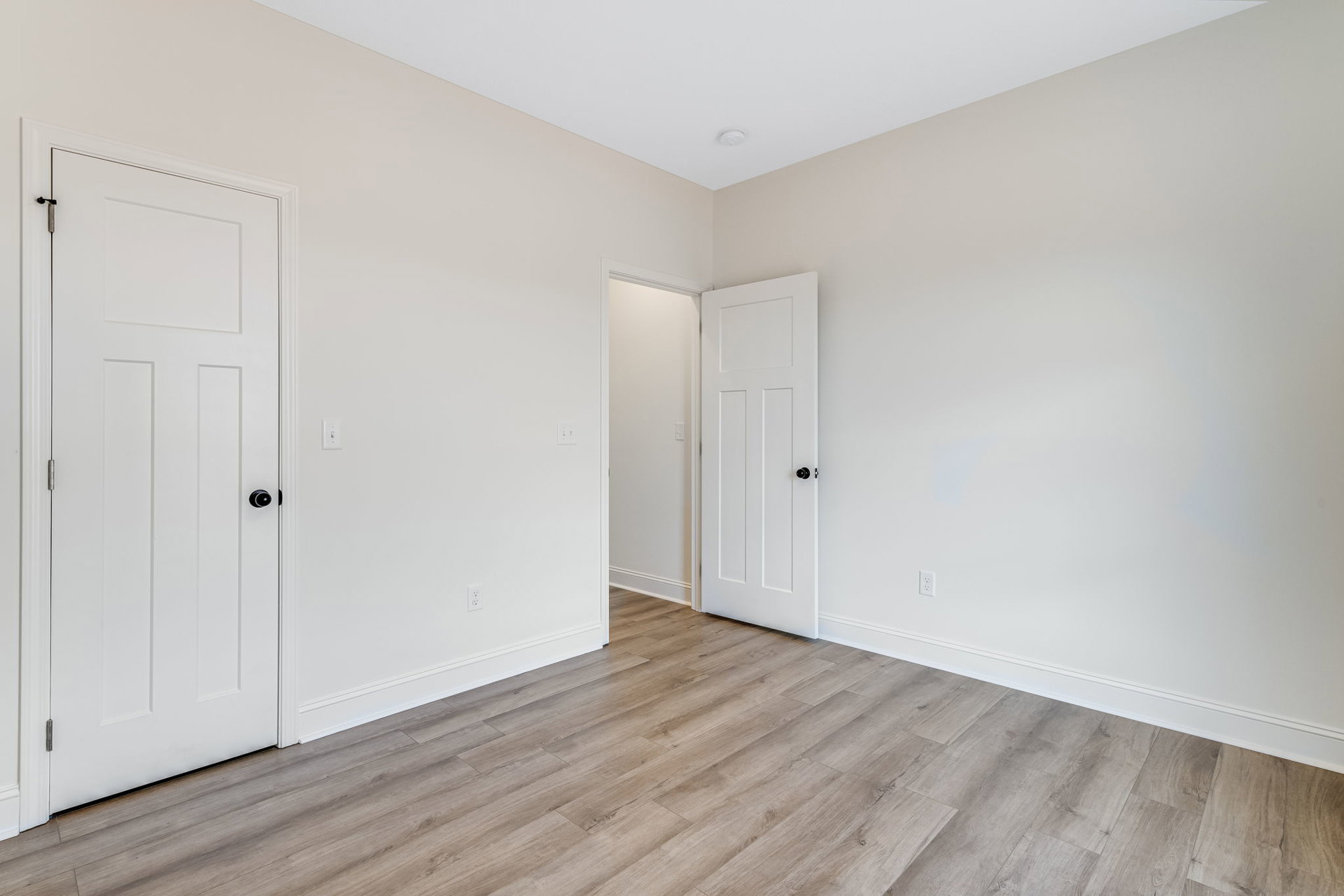 White walls and ceiling, wood laminate flooring, white door with black knob, light switch on wall, smoke detector mounted on ceiling