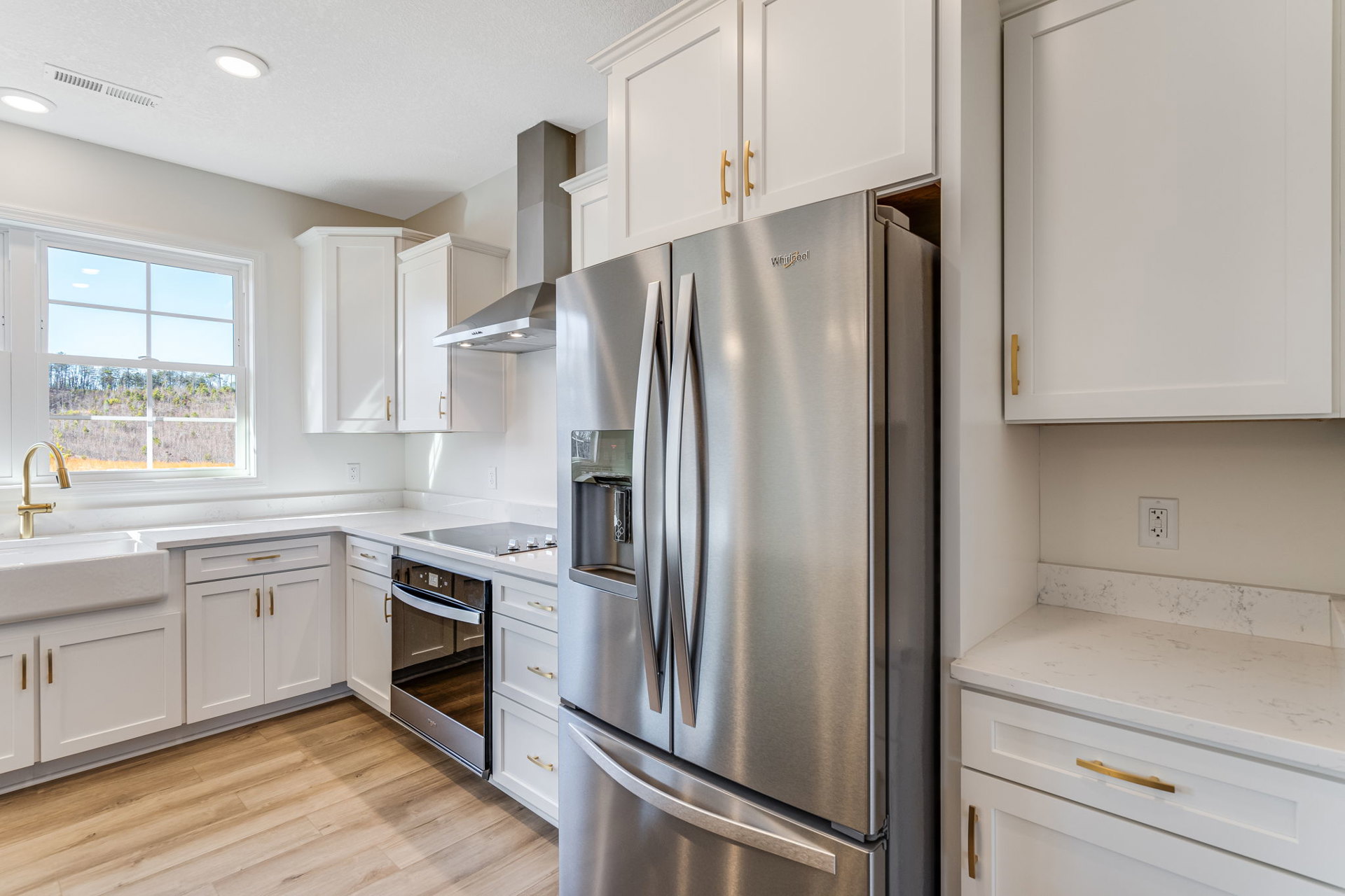 Modern kitchen featuring stainless steel refrigerator and oven, white cabinetry, black switch wall outlet, gold-handled white door, and window overlooking forest scenery