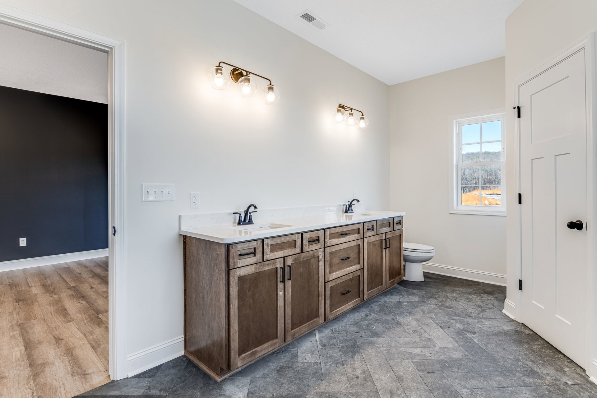 Bathroom with double sink vanity, black faucets, white toilet, tile floor, clear glass globe light fixture, window overlooking forest.