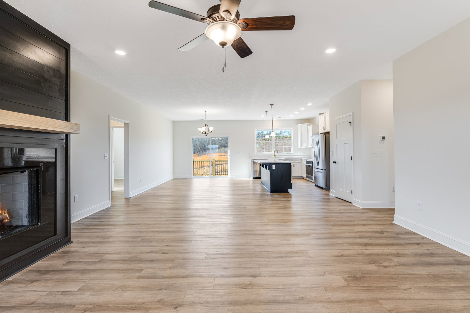 Open concept living area with wood flooring, ceiling fan with light fixture, black kitchen counters, stainless steel refrigerator, fireplace with active fire, and window