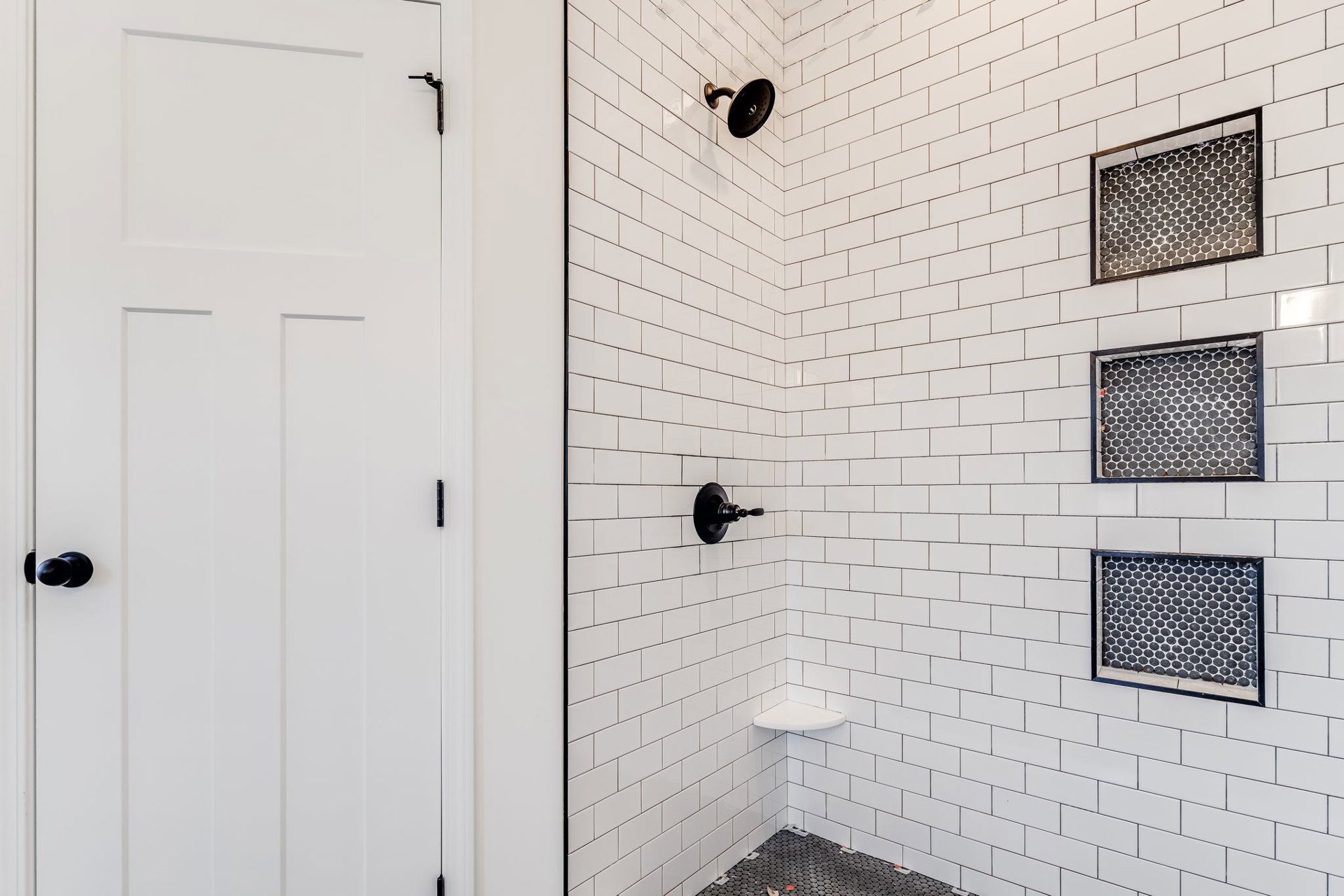 Shower with white tile walls, black and white tile floor featuring a black border, black knob on tile wall, white door with black handle
