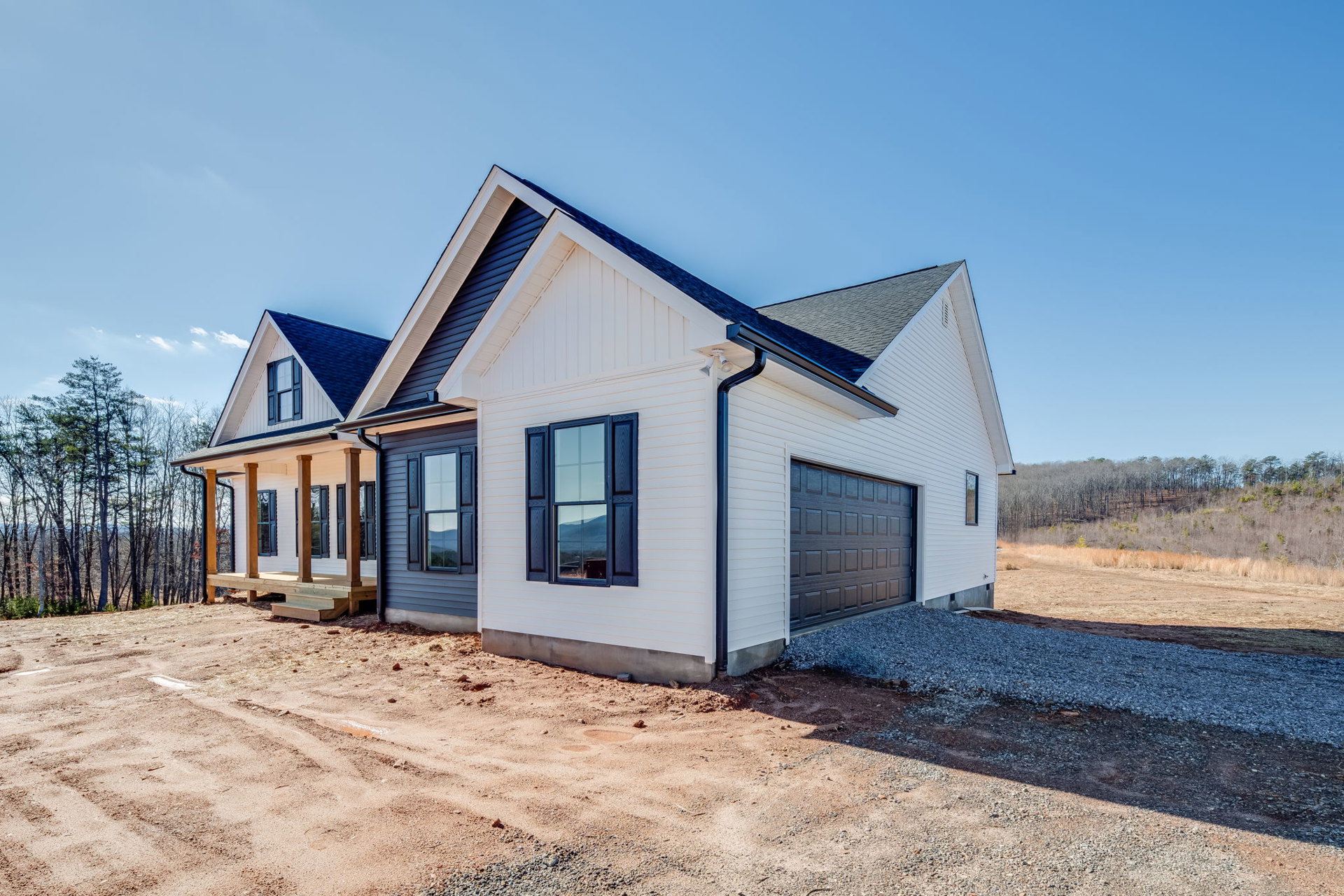 Two-story house with attached garage, covered porch, dirt driveway, large windows, and mountain view in the background, surrounded by trees and natural landscaping.
