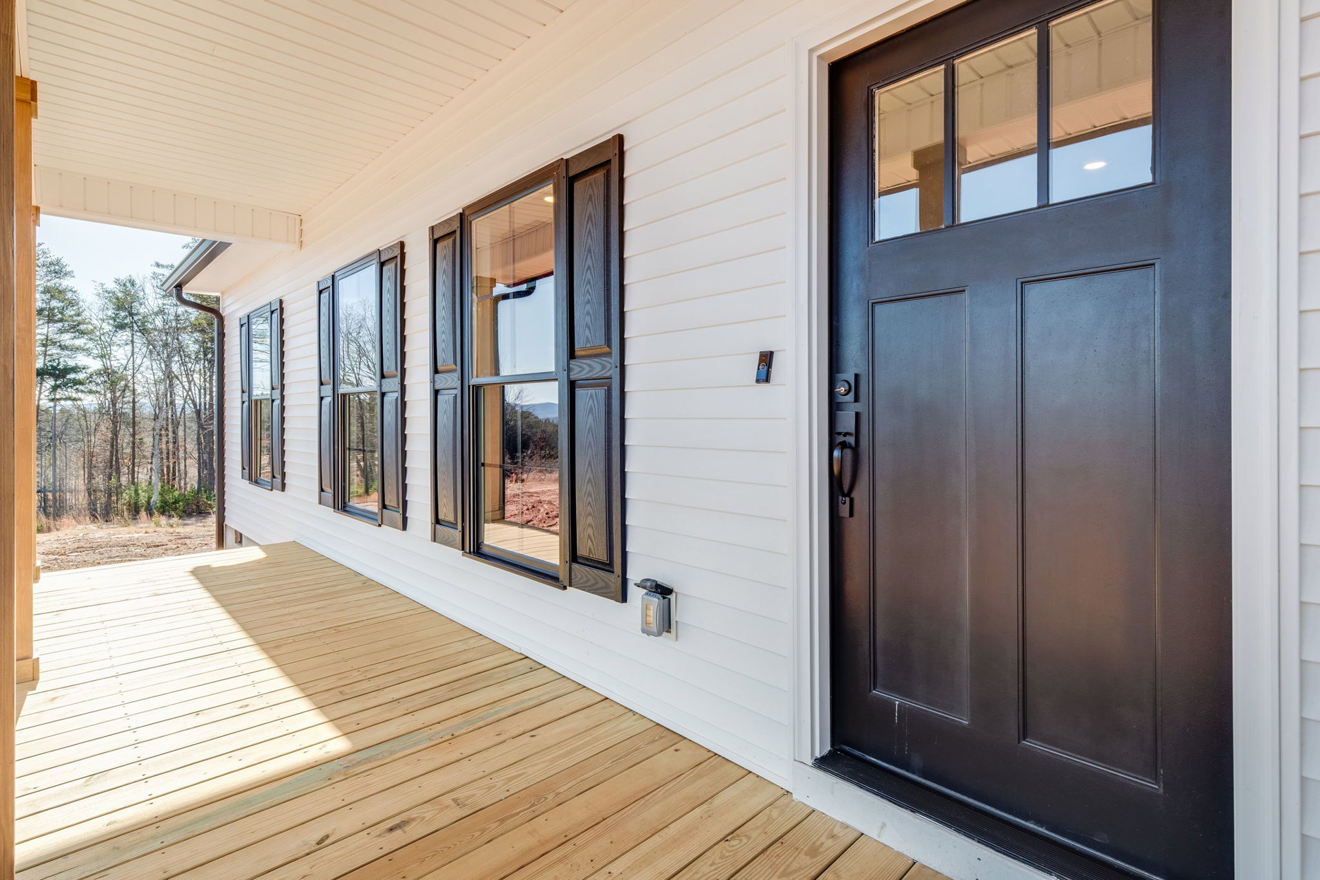 White siding exterior with black front door, large windows reflecting trees, covered porch, and wood flooring