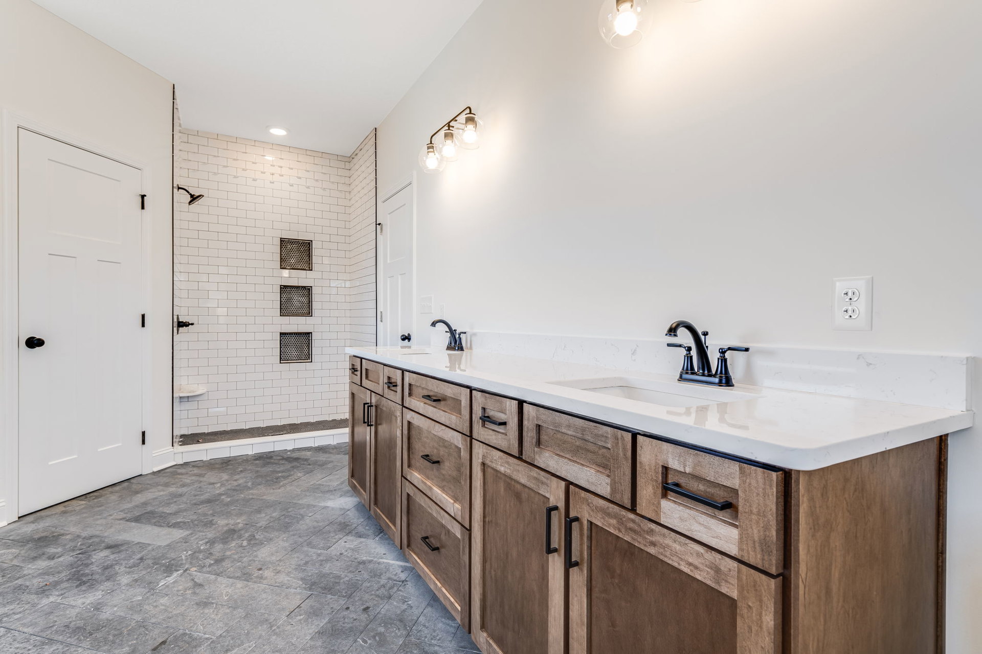 Bathroom with double sink vanity, white countertop, black faucets, glass-enclosed shower with tile walls, square window with grid, white door with black knob, visible electrical
