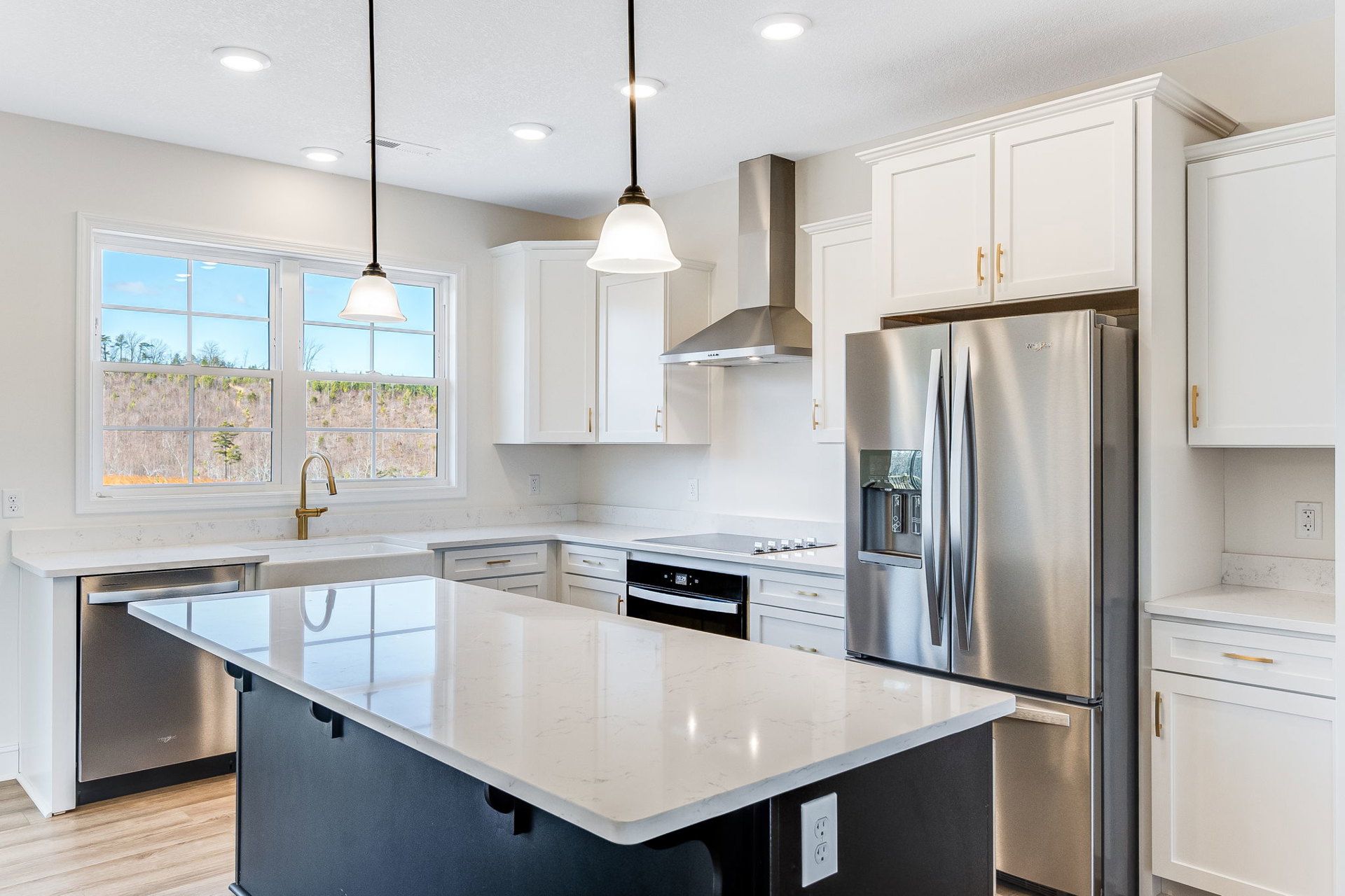 Spacious kitchen featuring a large white island with built-in sink, stainless steel refrigerator, white cabinetry, black and white oven, and window overlooking trees