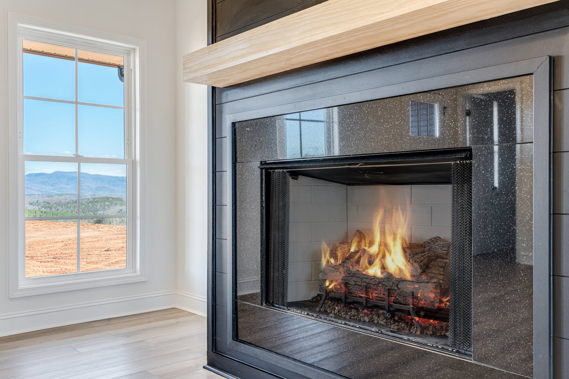 Stone fireplace with wood mantel, fire burning, large window framing mountain view, hardwood floors, neutral walls