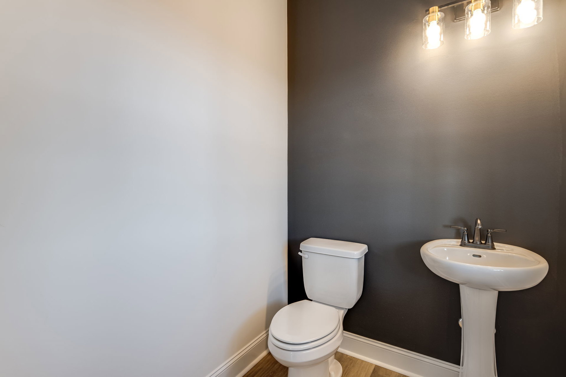 White pedestal sink with matte black faucet beside a white toilet, both set against smooth white tile walls in a minimalist bathroom.