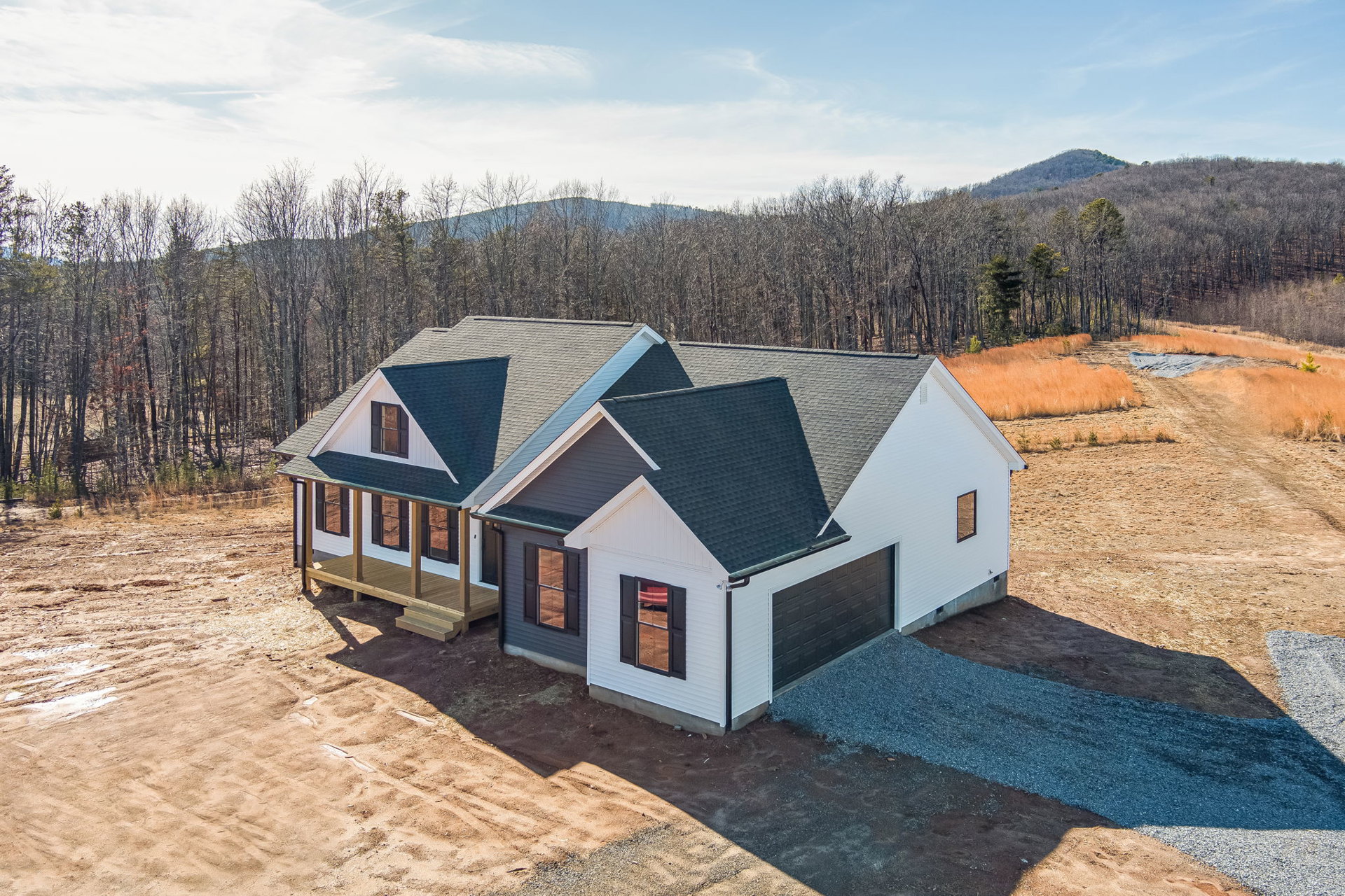 Two-story home with gray siding, covered front porch, attached garage, expansive concrete driveway, large windows reflecting red blanket, surrounded by trees under partly cloudy