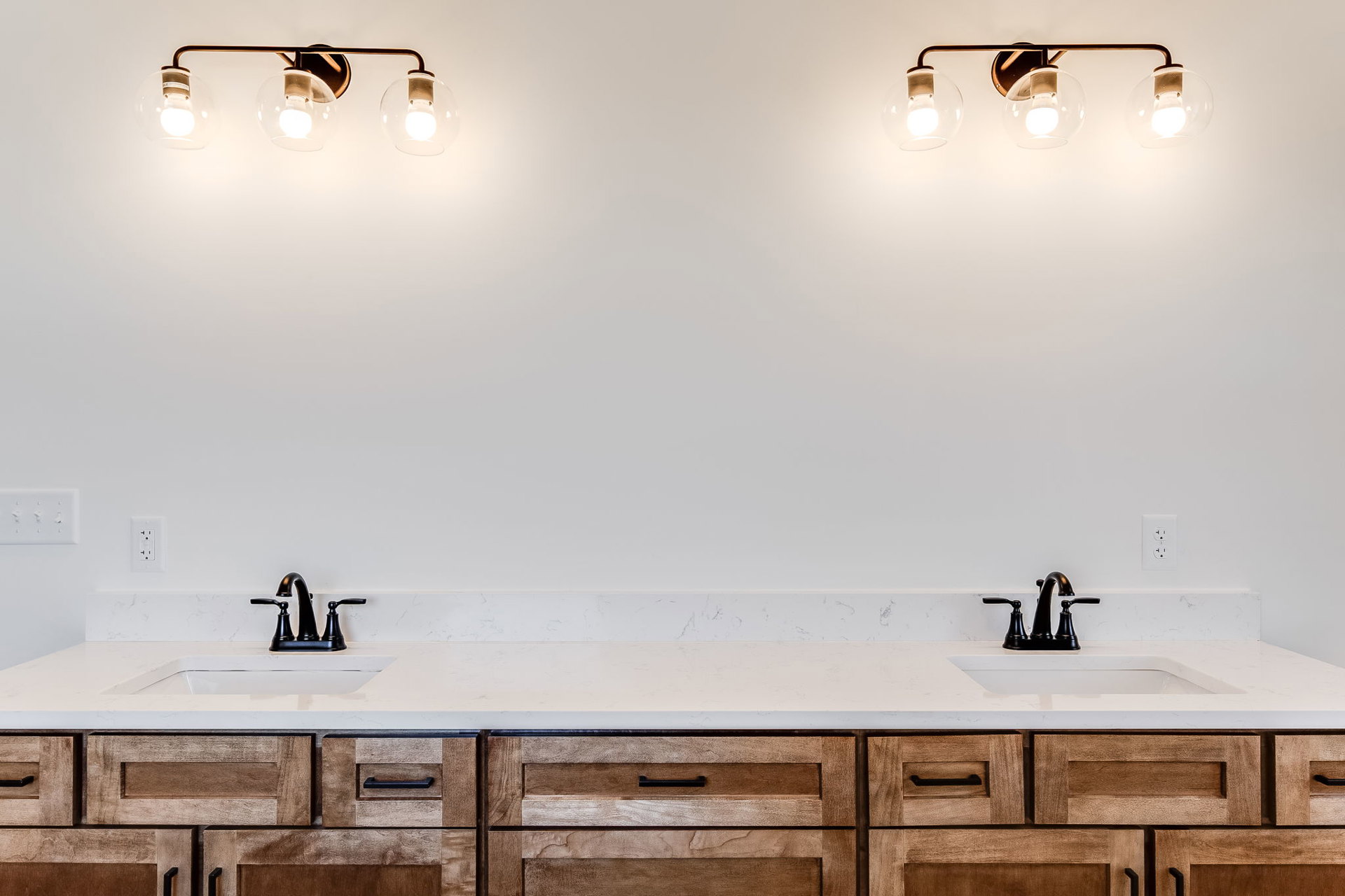 Bathroom with white painted walls, dual undermount sinks set in a light stone countertop, chrome faucets, white cabinetry, modern light fixtures with exposed clear bulbs, and