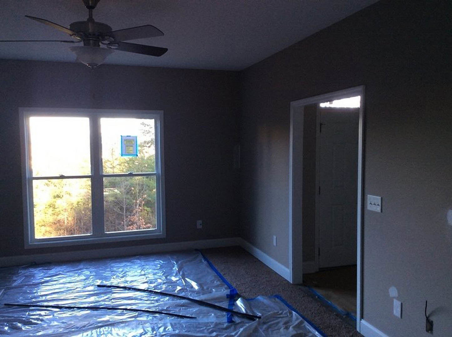 Neutral-toned room with a ceiling fan and light fixture, single window with white trim, smooth walls, and visible electrical outlet