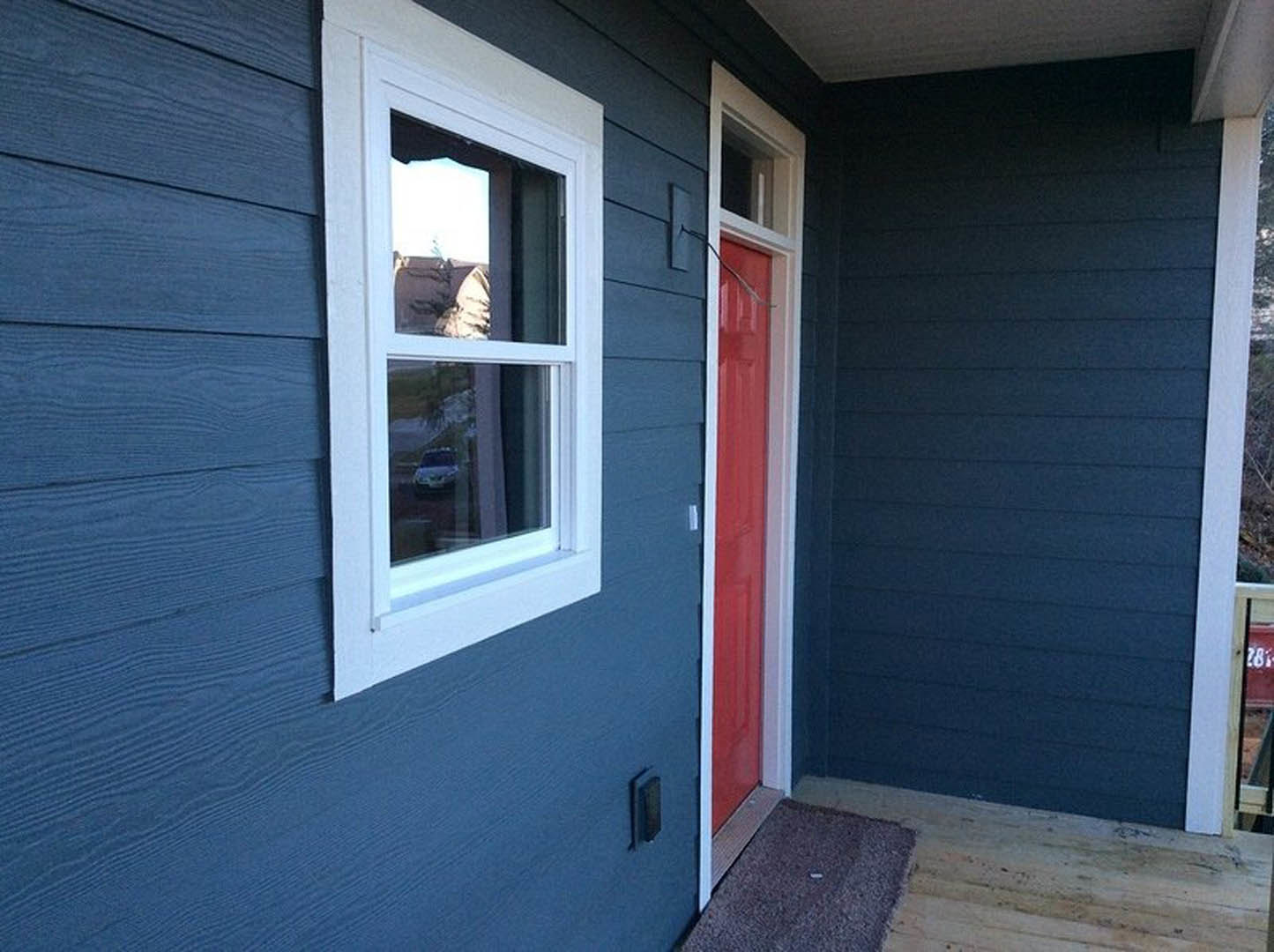 Blue siding exterior with white trim, red front door, large windows reflecting a parked car, concrete walkway, landscaped front yard