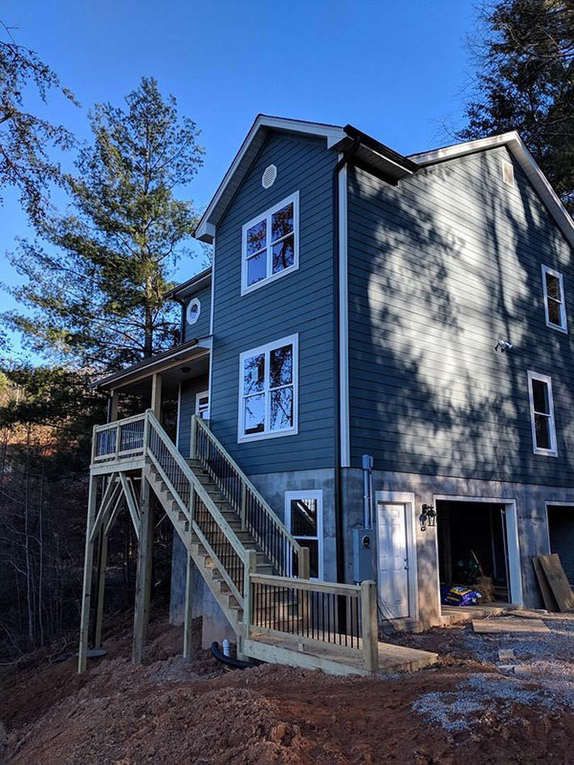 Wooden exterior staircase leading to a porch, white-framed window reflecting trees, dark entryway with open door, surrounded by outdoor greenery.
