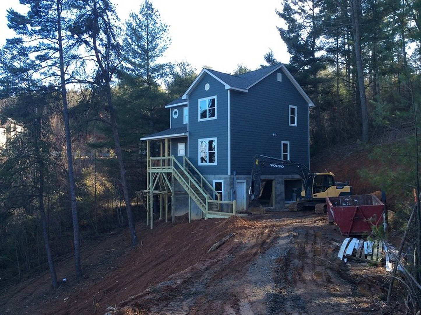 Wood-framed house under construction on a hill, exterior staircase, crane, red shipping container, white-framed window, surrounding trees and sky