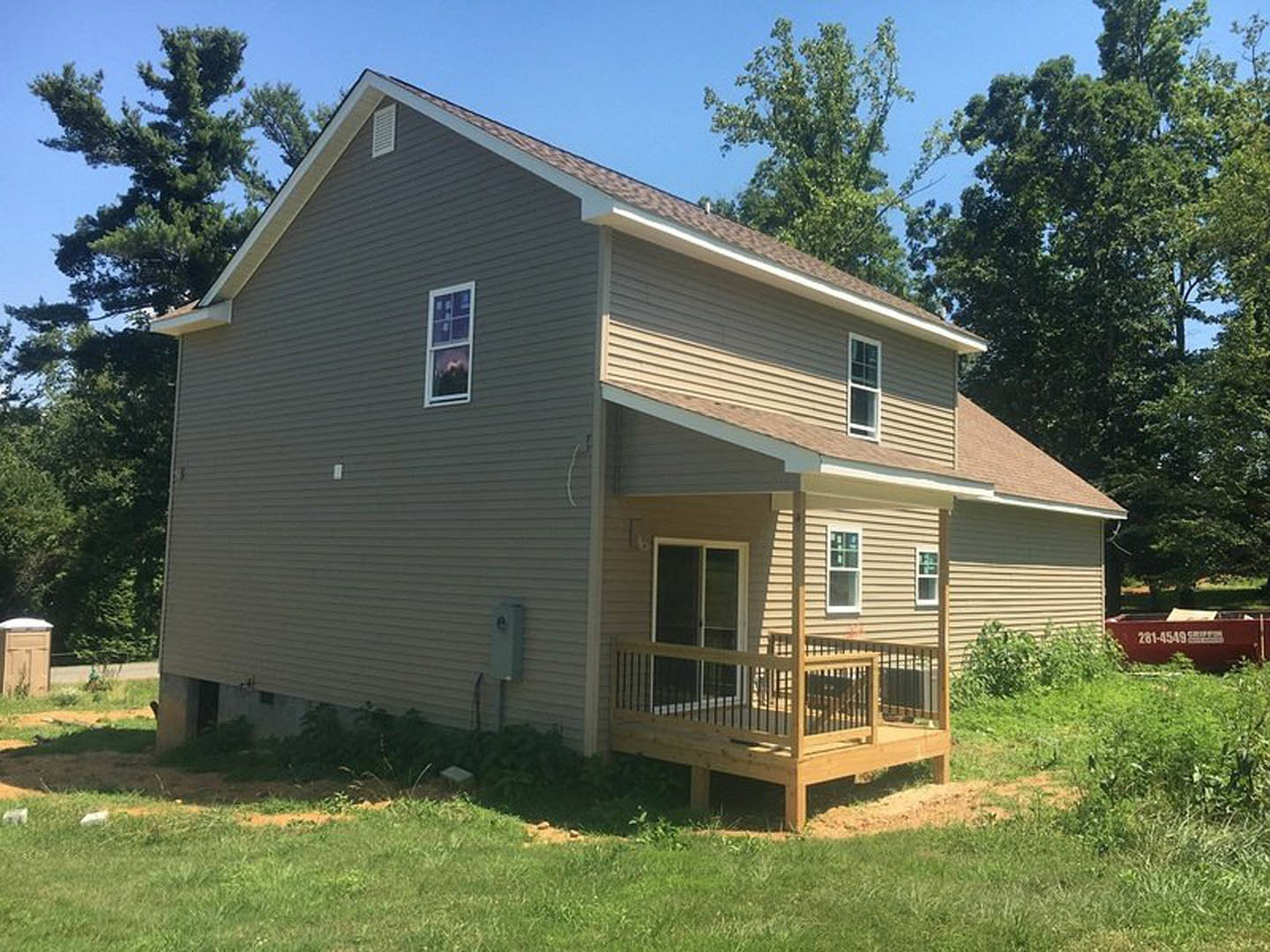 Two-story home with light siding, covered front porch, wooden deck with railing, large windows, manicured lawn, and surrounding plants.