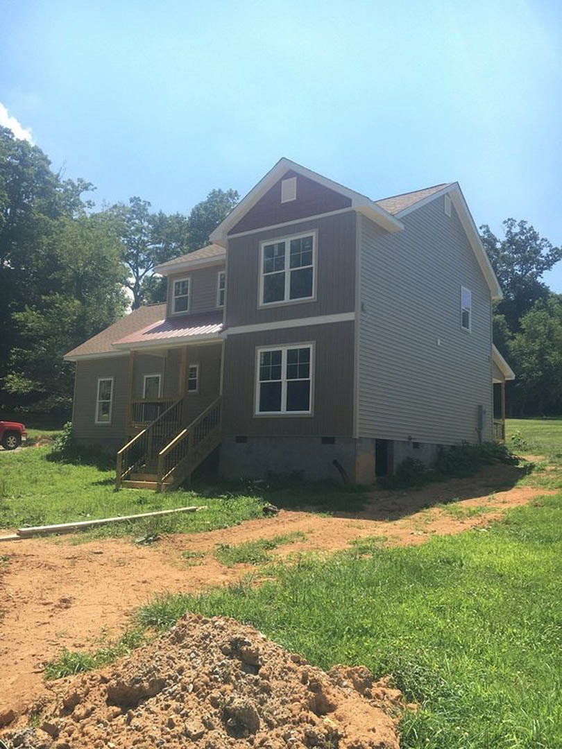 Two-story house with white-framed windows, wooden staircase leading to entrance, dirt path and pile of soil on grassy yard, surrounded by trees under blue sky