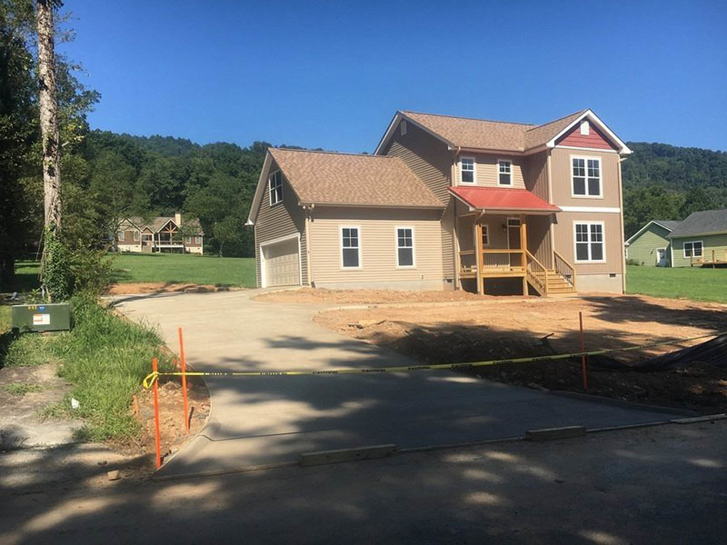 Two-story house with red roof, front porch, multiple windows, concrete driveway, green lawn, and trees under a clear blue sky