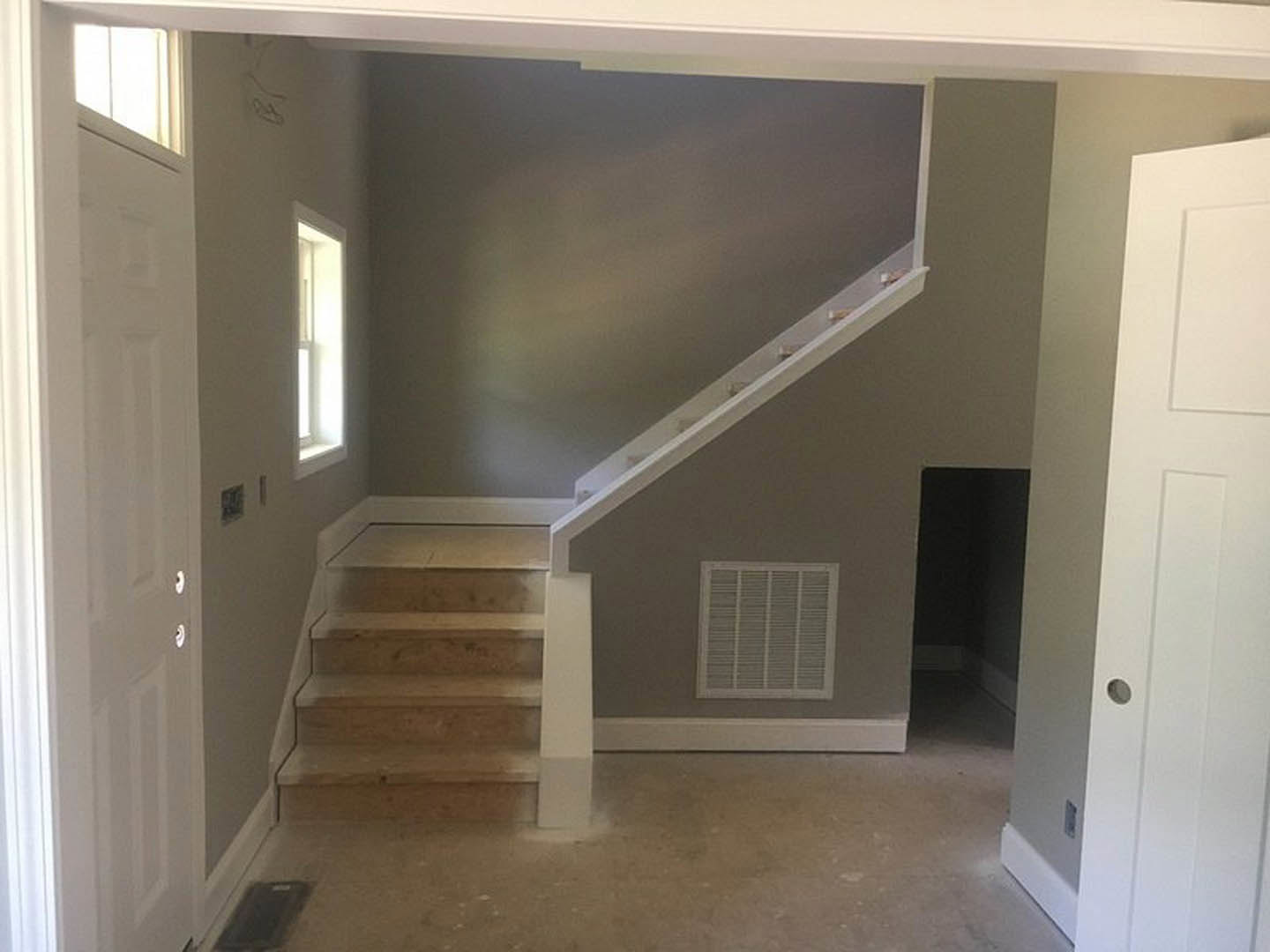 Wood staircase with white risers and molding, adjacent to a vented wall and white door, sunlight streaming through nearby window onto light-colored flooring and plaster walls