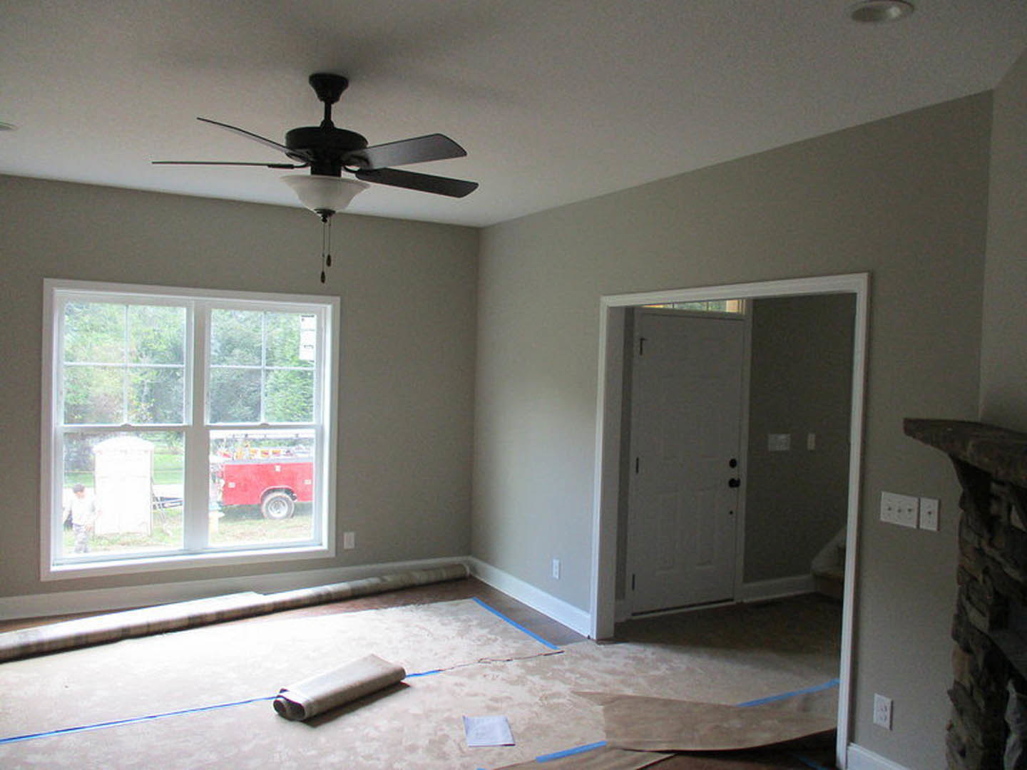 Bedroom with beige carpet flooring, white walls, ceiling fan with light fixture, large window overlooking driveway with red truck and ladder visible outside