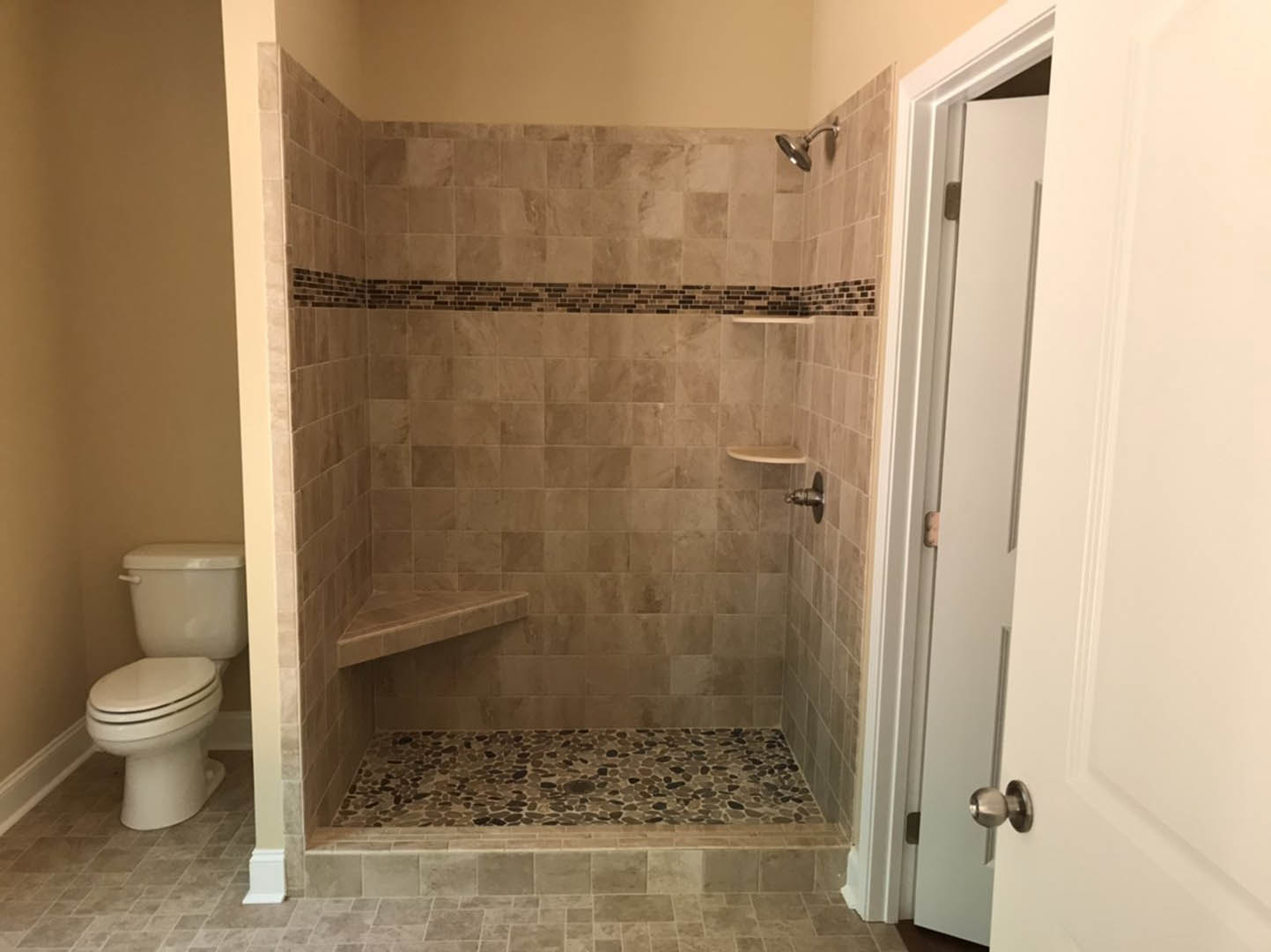 Walk-in shower with gray tile floor and walls, white toilet with closed lid, white paneled door with silver door knob, bathroom interior with neutral finishes