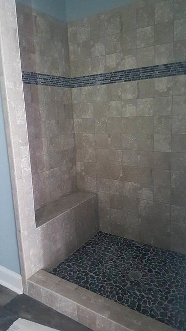 Tiled shower featuring a built-in bench, linear shower drain, grey and black tile flooring, and a brick accent wall.