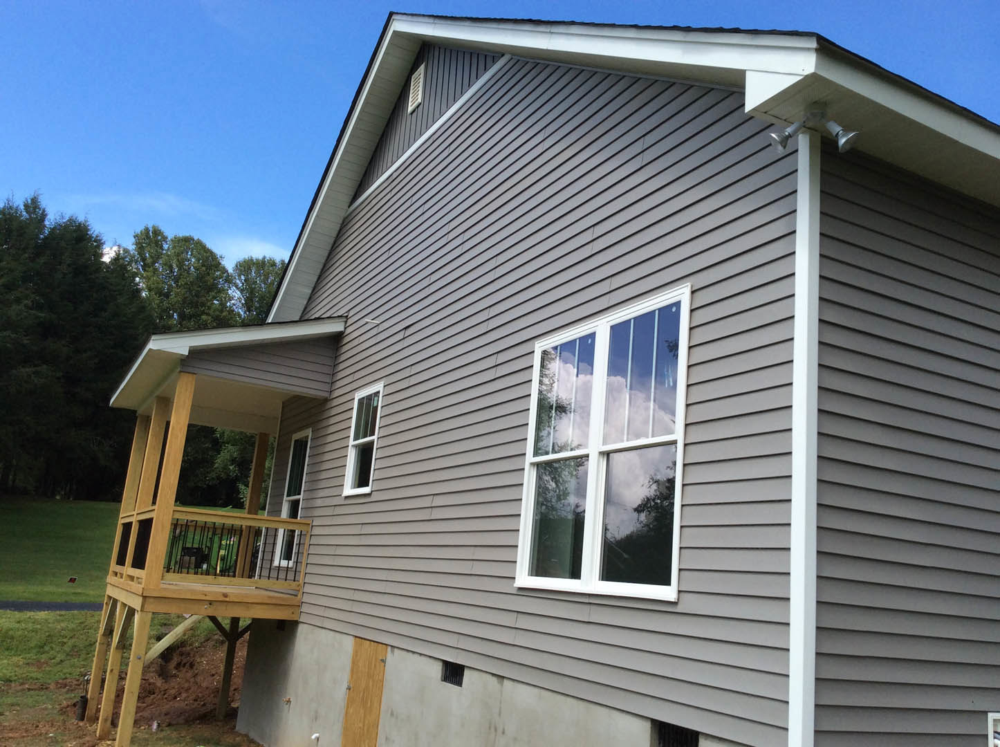 Wooden deck and covered porch with white siding, large windows, and gabled roof surrounded by mature trees